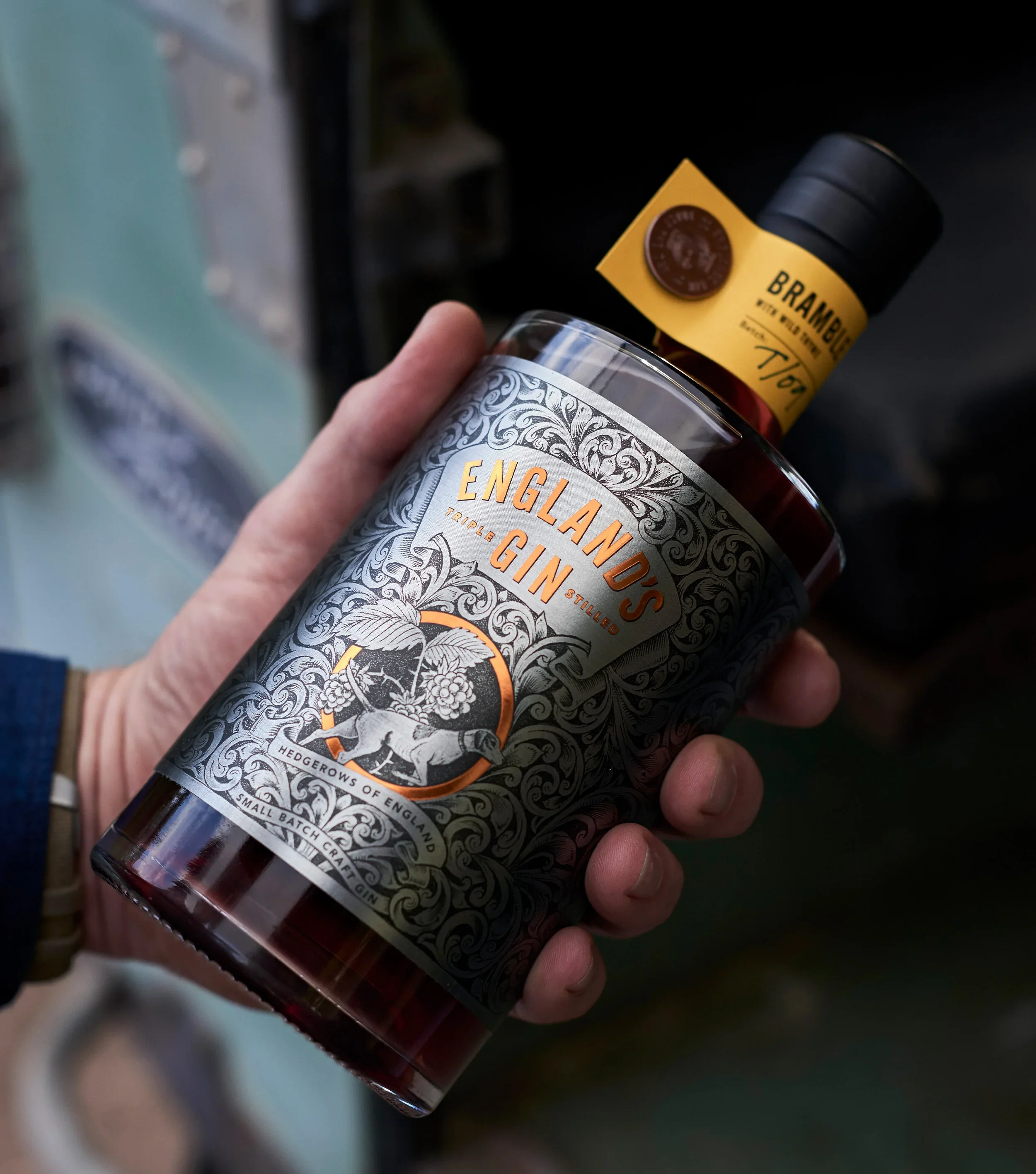 Close-up of a hand holding a bottle of English gin with a decorative label and a yellow tag attached to the neck of the bottle.