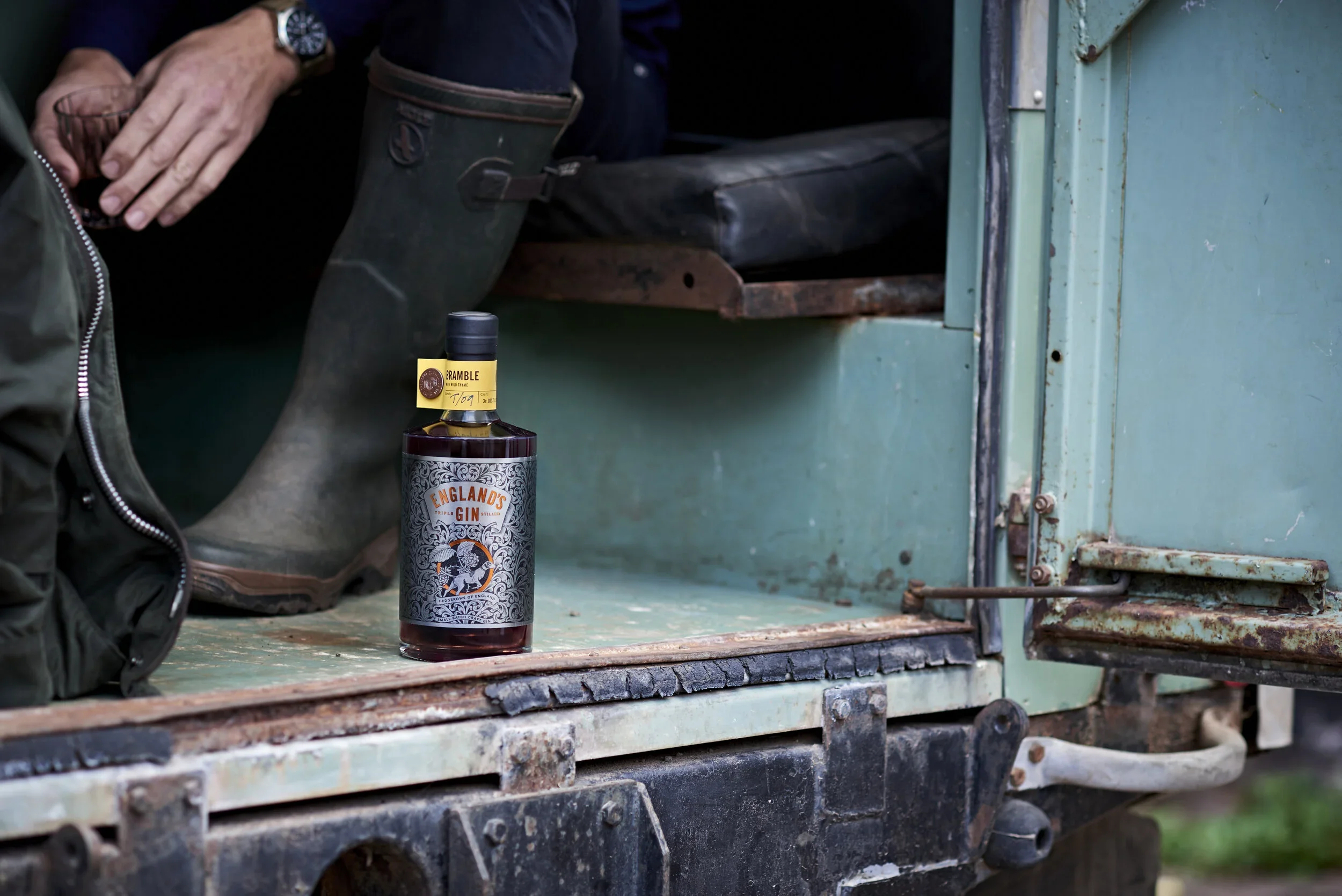 A bottle of England's Gin placed on a rusty, weathered teal-colored metal surface next to a person wearing a watch and dark clothing, with a black bag and a rugged environment.