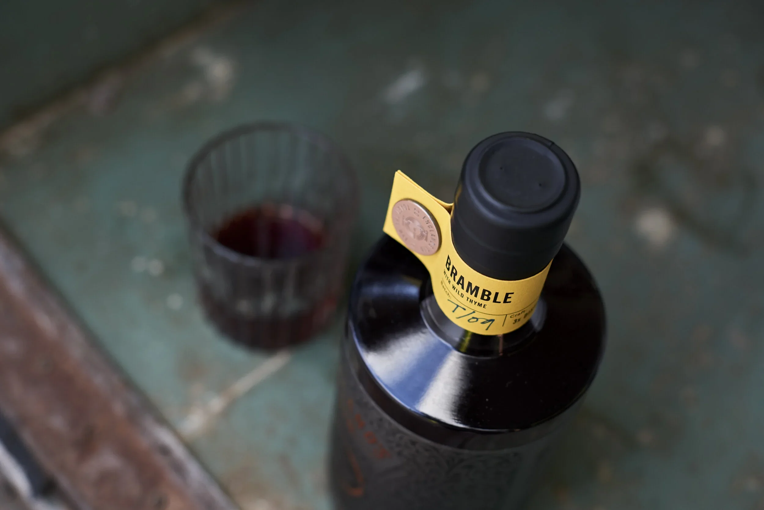 A bottle of Bramble with Wild Thyme lying on a black surface, viewed from above. There is a glass filled with red liquid in the foreground, seen from above. The setting appears to be a kitchen countertop.