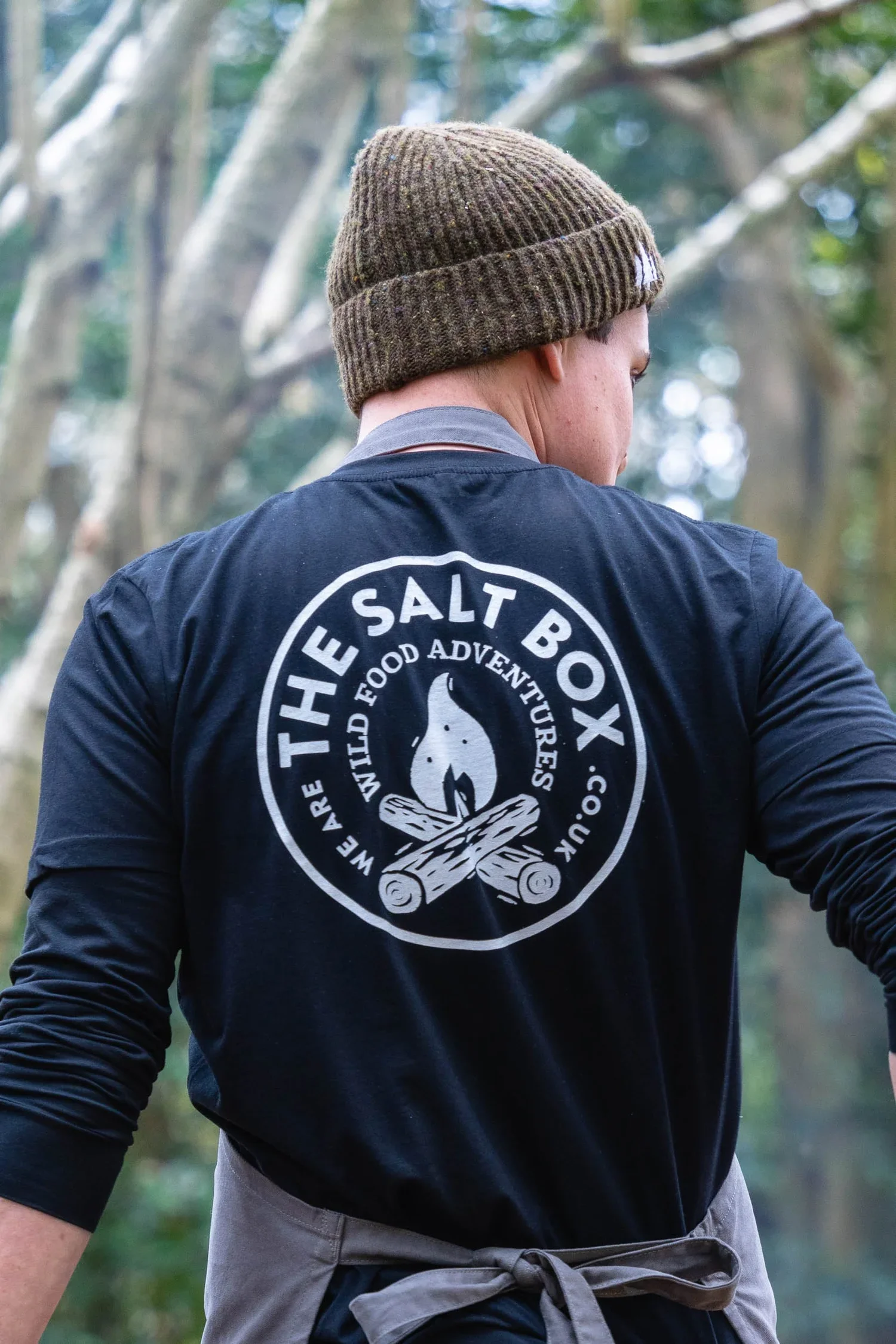 A man wearing a brown knit beanie and a black jacket with a white circular logo on the back, standing outdoors among trees with emerald green leaves, with part of his face and body visible.