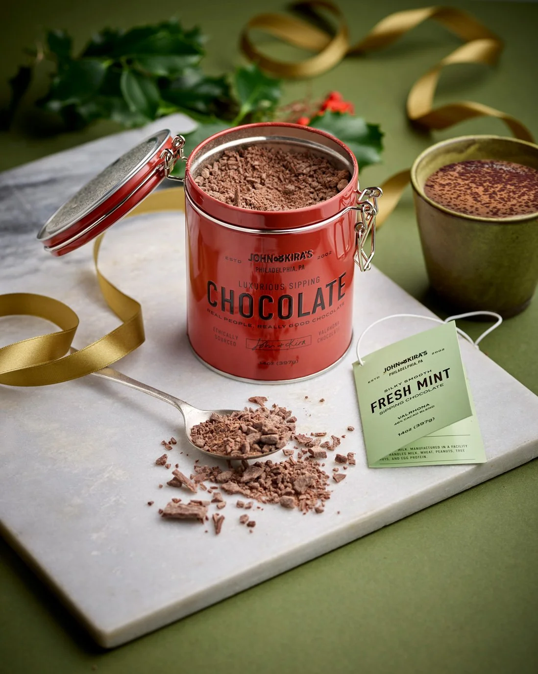 Open red chocolate tin with crushed chocolate inside, on white marble surface with a spoonful of crushed chocolate, green leaves, a gold ribbon, and a cup of hot chocolate in the background.