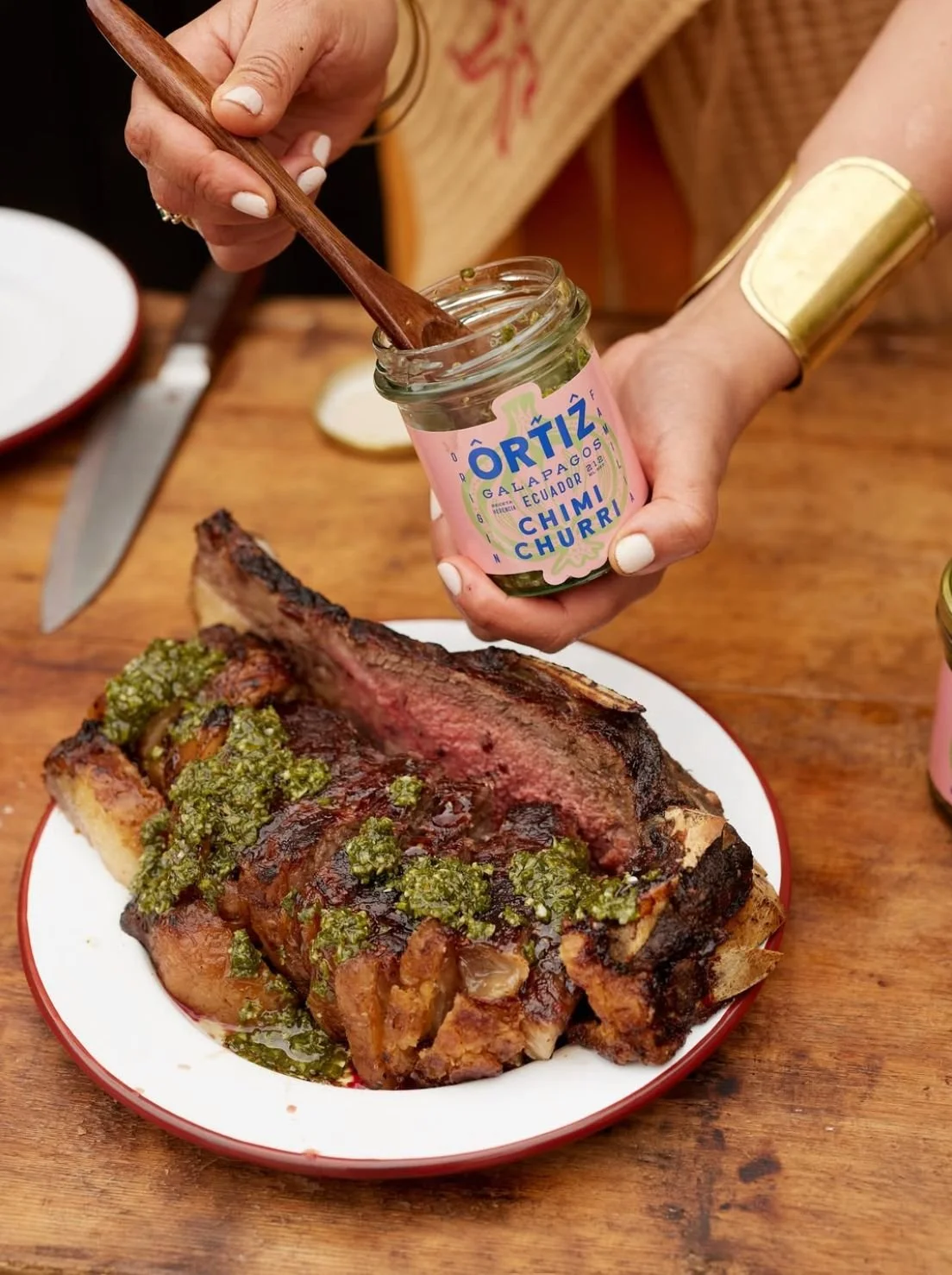A person spreading green chimichurri sauce on a grilled steak, which is served on a white and red plate on a wooden table.
