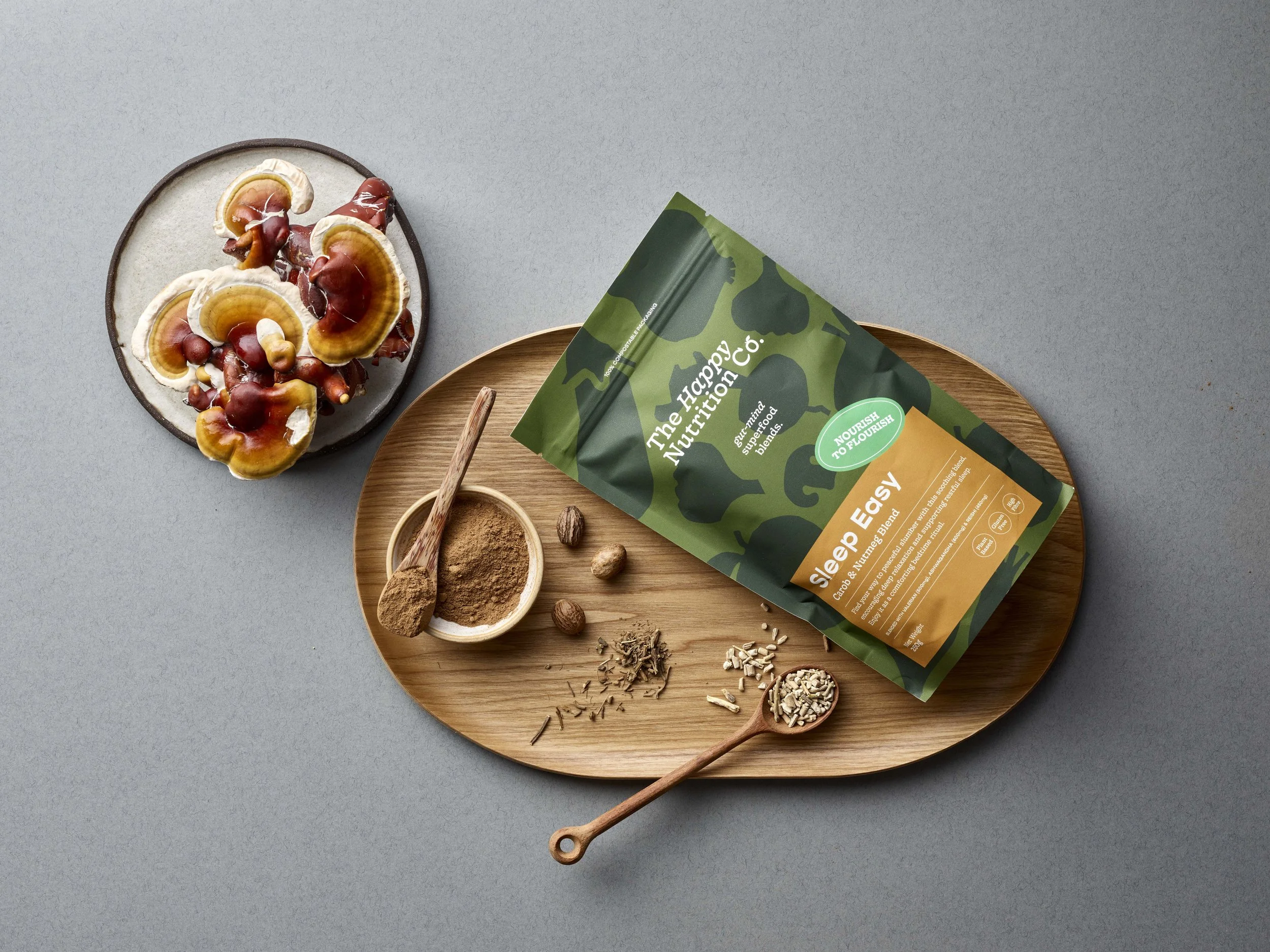 Flat lay of a gray surface with a ceramic plate containing reishi mushrooms, a wooden tray with a green health food bag and various spices and herbs, and small wooden spoons with powders and seeds.