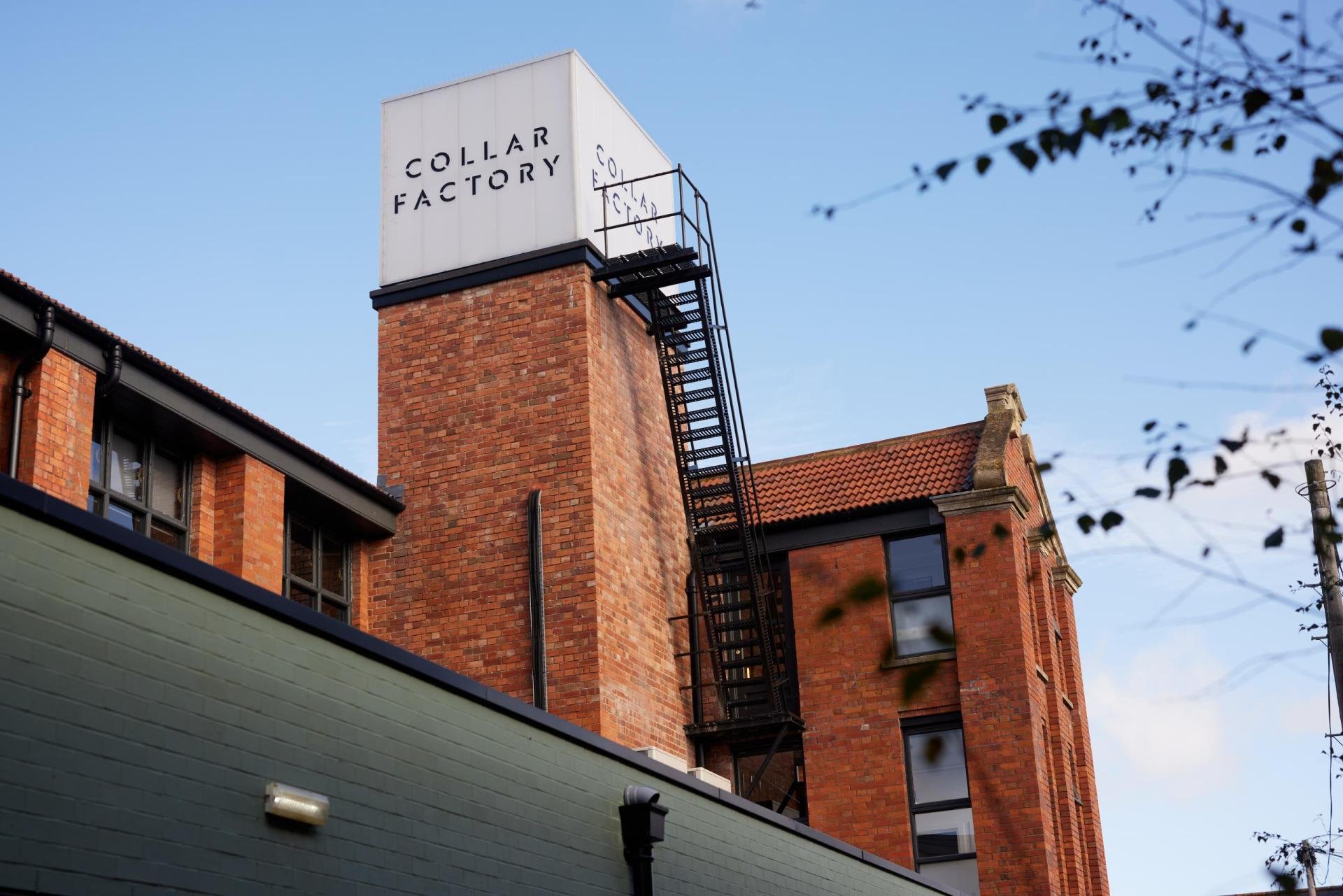 A brick building with multiple windows, a fire escape, and a large white sign on top that reads 'COLLAR FACTORY,' set against a blue sky.