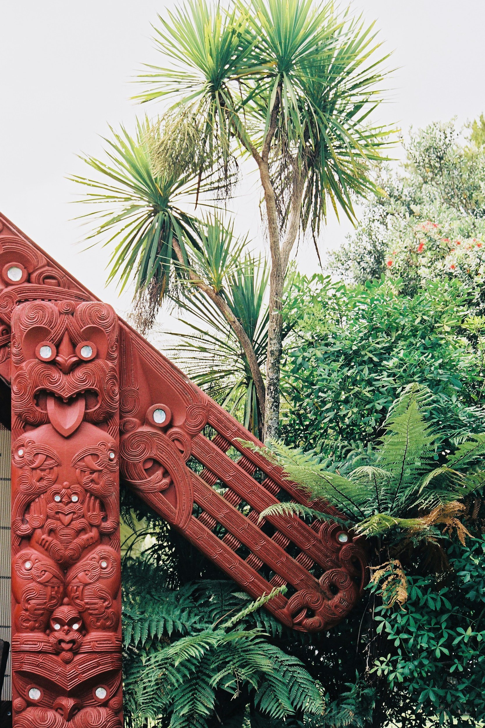 Red carved Maori-style tiki sculpture in a lush green garden with tall palm trees and dense foliage.