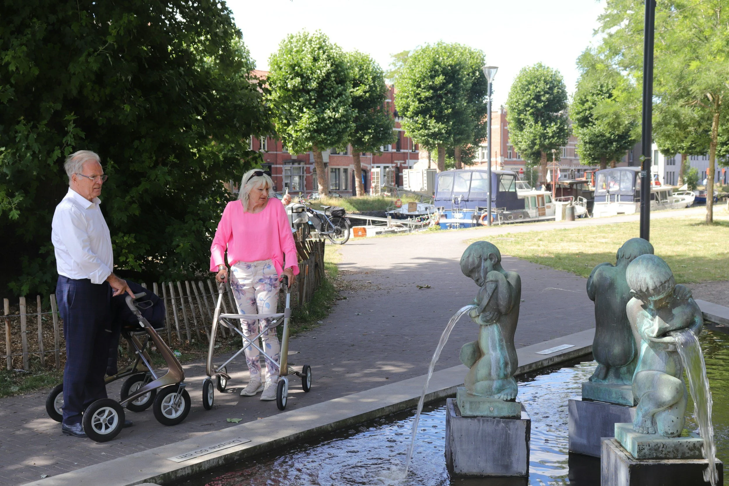 Two people at a park newar a water fountain, one person is weairng a white shirt and navy pants with glasses, the other is wearing a pink top tucked into light coloured floral patterned pants. They both have mobility walkers assisting them.