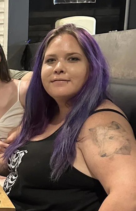 A woman with long purple and blue hair sitting in a restaurant or cafe, wearing a black tank top with a lion tattoo on her upper arm.