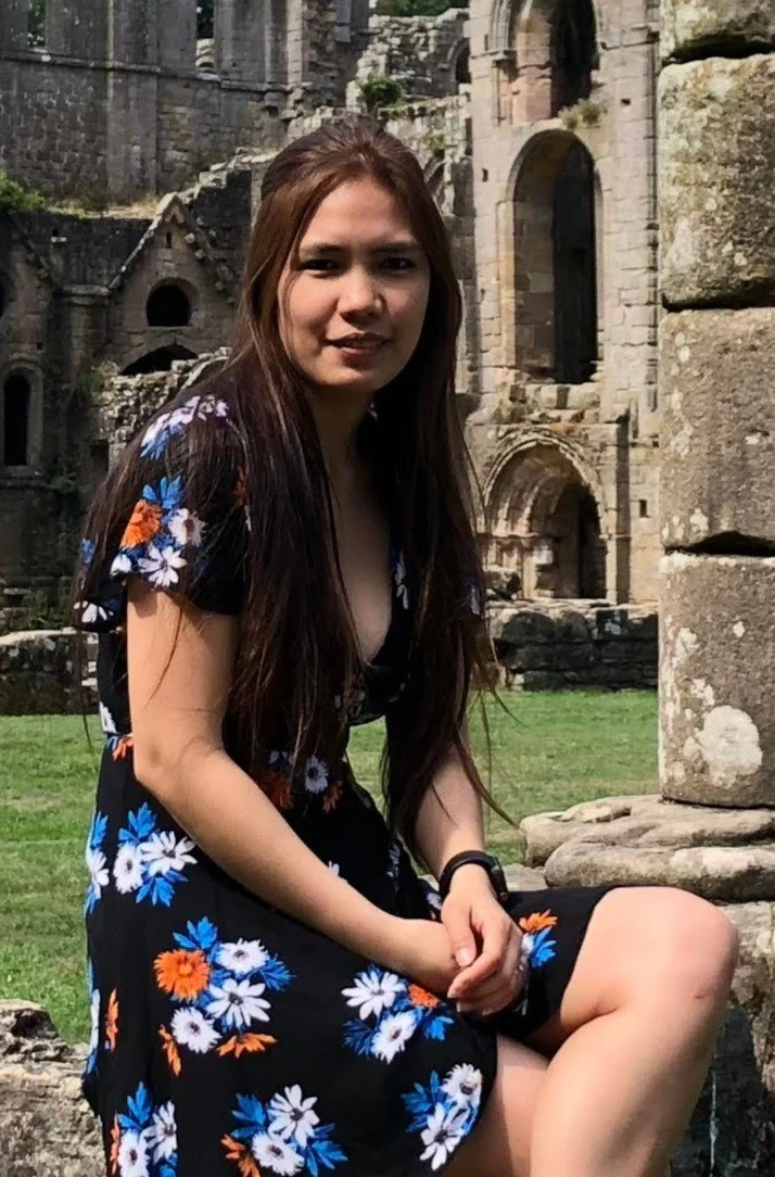 A young woman with long brown hair sitting outdoors near ancient stone ruins, wearing a black floral dress with white, blue, and orange flowers.