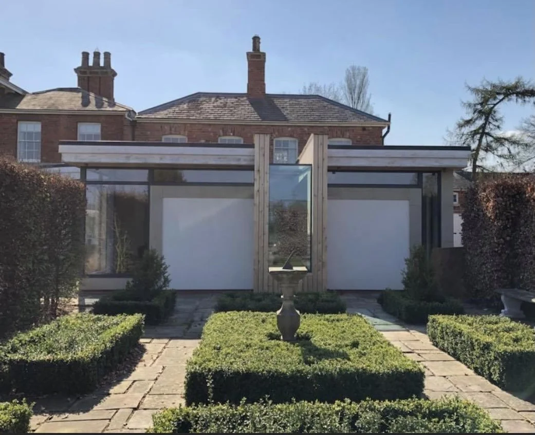 Modern greenhouse with glass walls and wooden accents, surrounded by neatly trimmed hedges and a stone pathway, with a house and trees in the background.