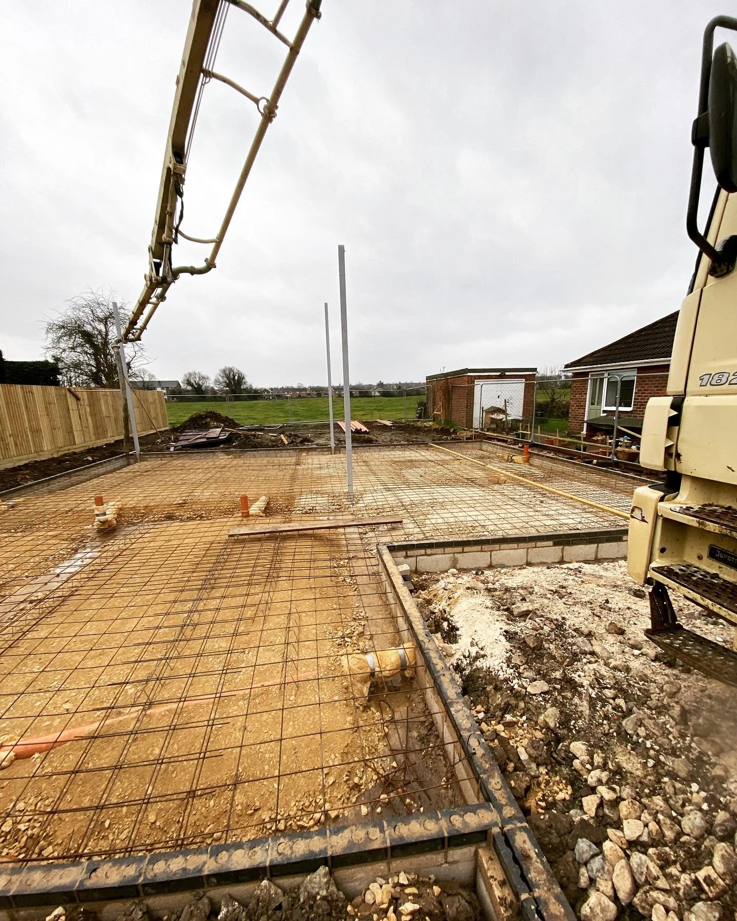 Construction site with a foundation and steel rebar, a concrete pump arm in the air, and neighboring houses and fields in the background.