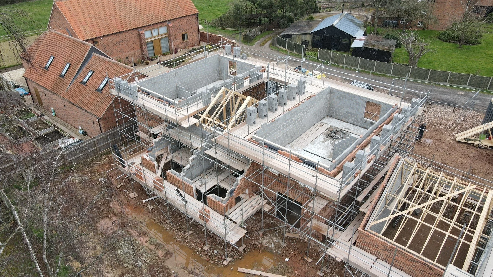 Aerial view of a house under construction with scaffolding, concrete block walls, and wooden framing, surrounded by neighboring houses and yards.