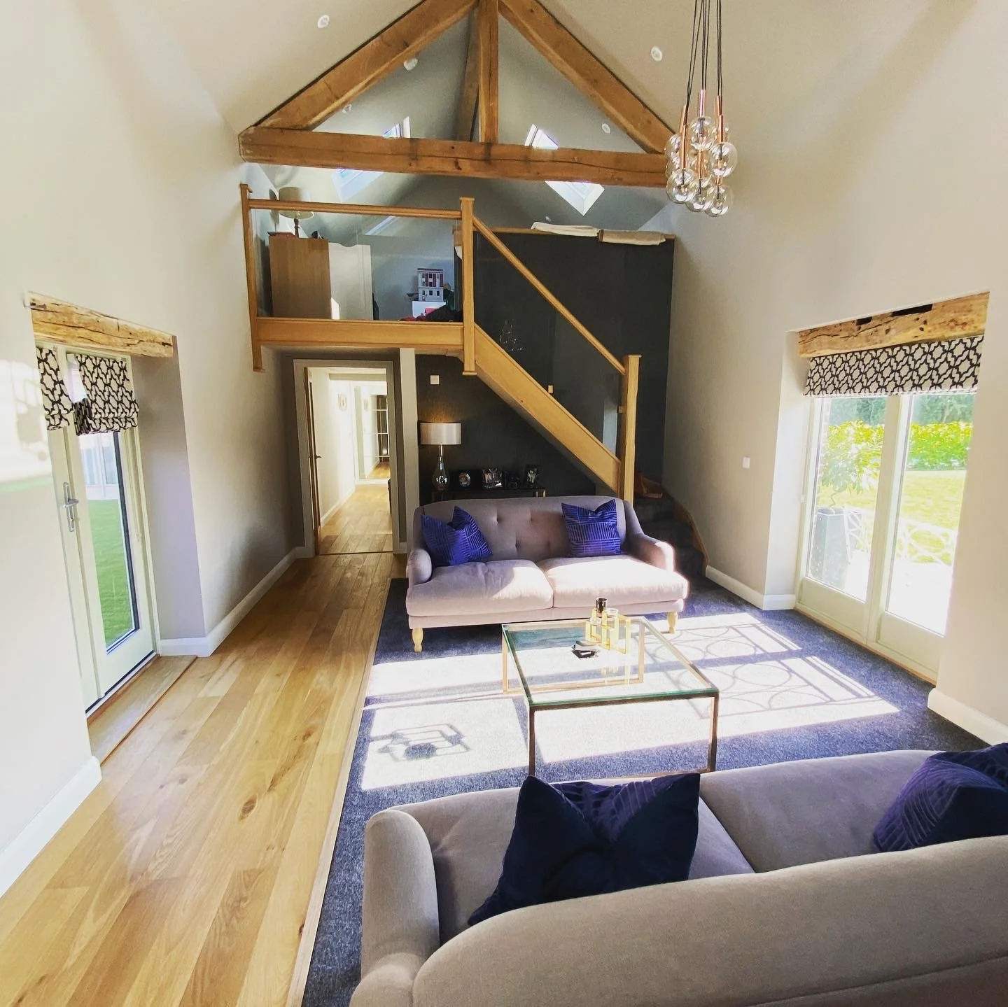 Interior of a living room with high vaulted ceiling and wooden beams. There are two sofas with purple pillows, a glass coffee table with decorative bottles, and a dark accent wall with a staircase made of wood leading to a mezzanine. Large windows an