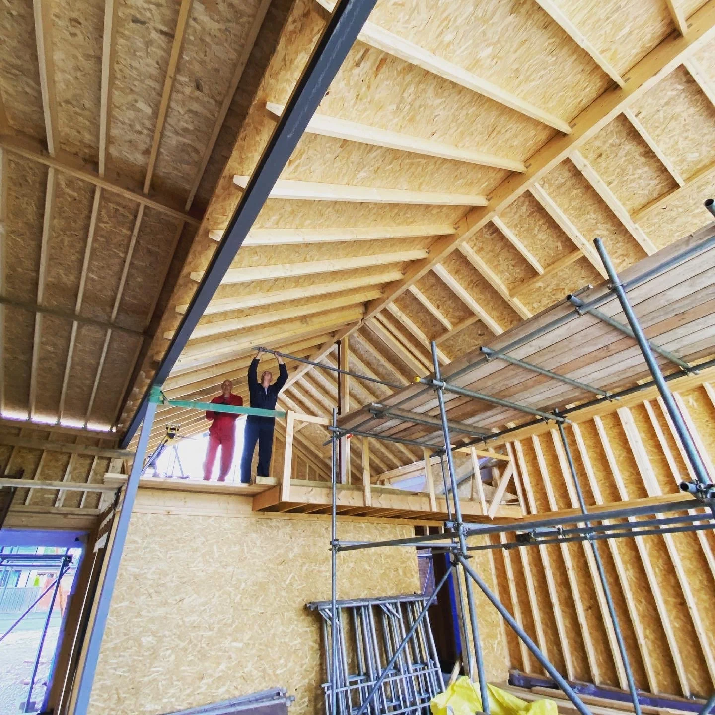 Two construction workers stand on a wooden platform inside a building under construction, surrounded by scaffoldings and exposed wooden framing.