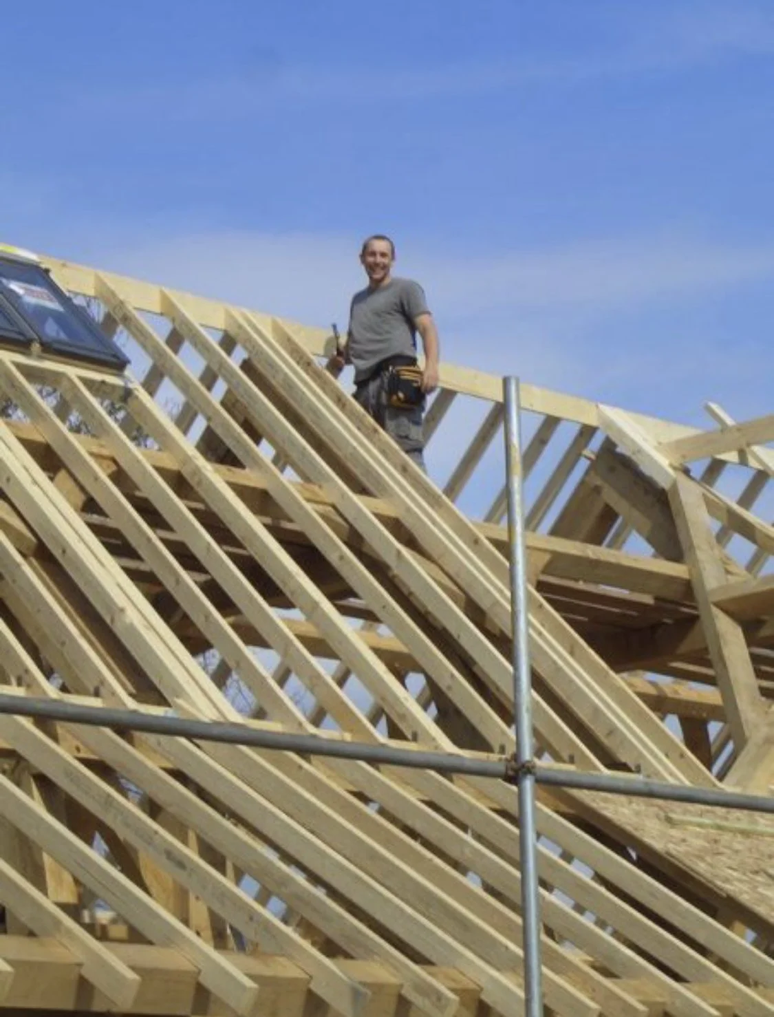 A man working on a roof under construction, standing on wooden framing with blue sky in the background.