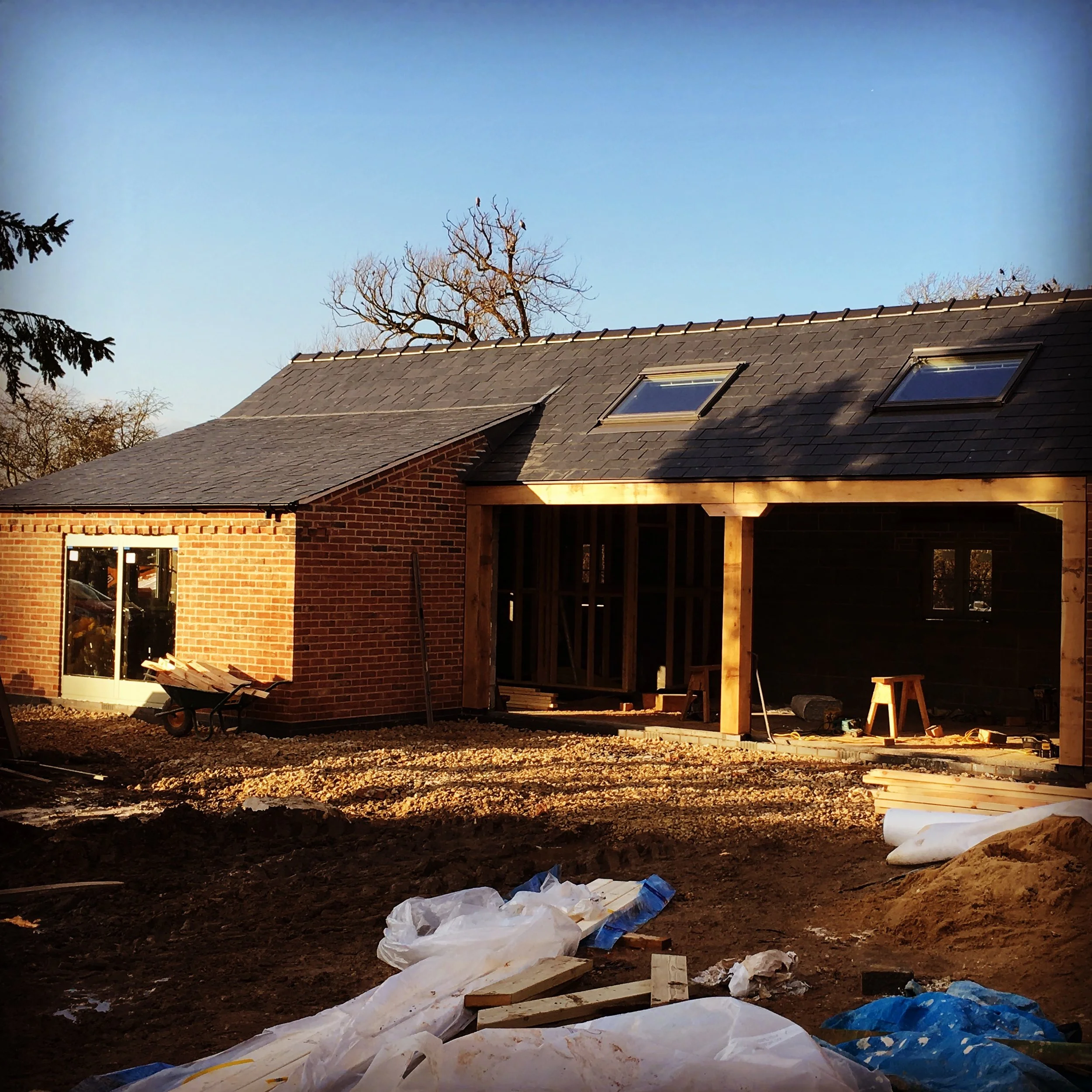 Construction of a house with brick and wooden framing, with partially completed walls and roof, construction materials and tools on site, and a clear blue sky in the background.