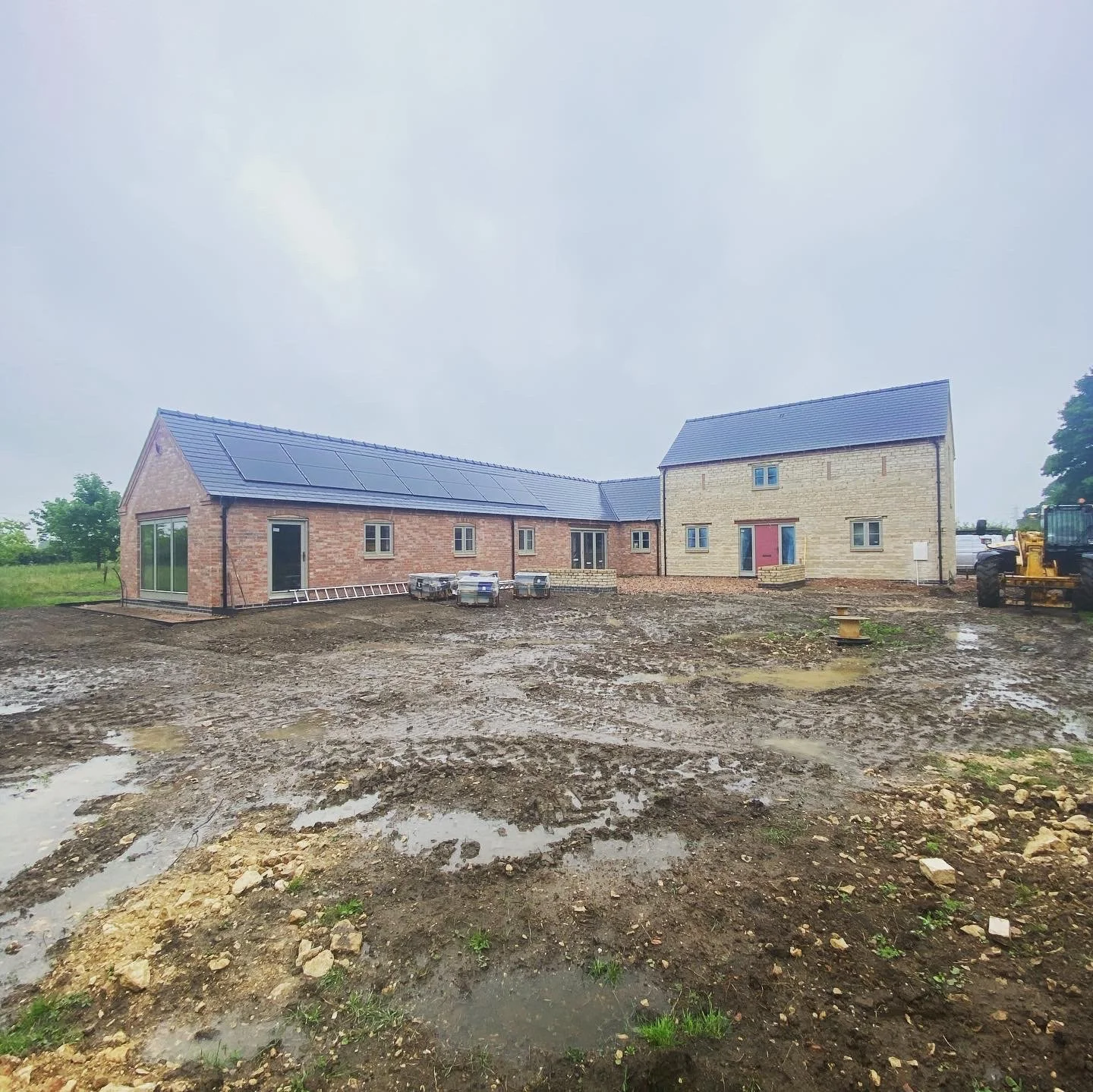 Under construction rural house with solar panels on roof, muddy ground with puddles, construction materials, and machinery, cloudy sky in background.