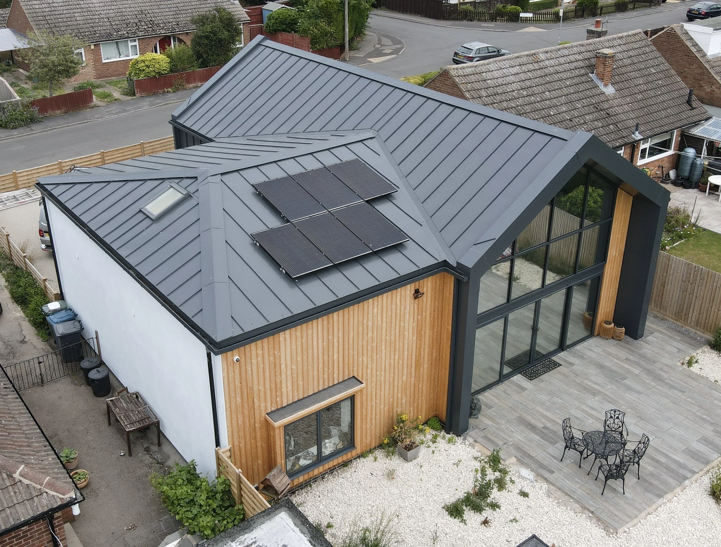 Modern house with metal roof, solar panels, large glass windows, wooden paneling on one side, a patio with black metal outdoor furniture, and a gravel yard.