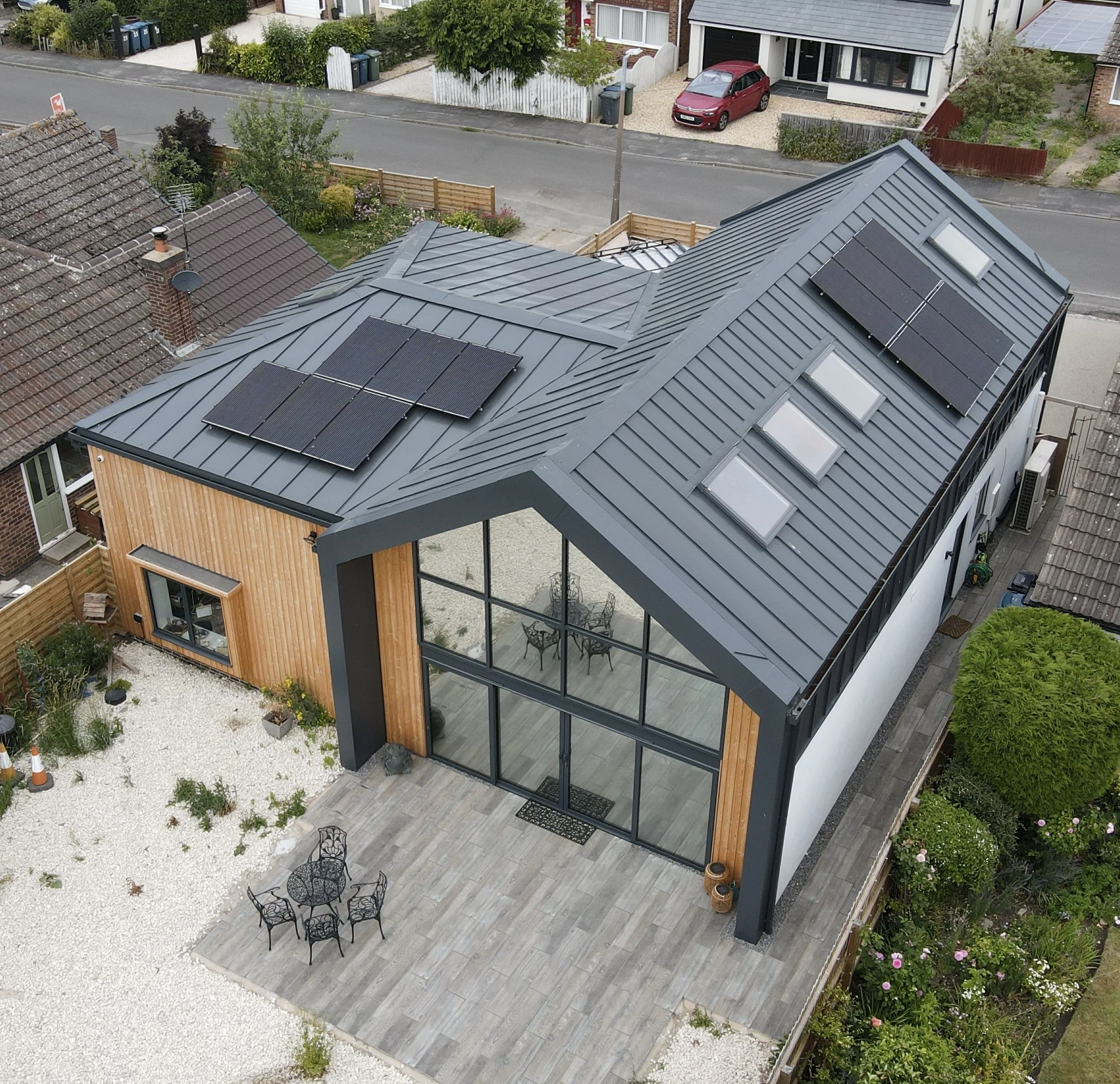 A modern house with black metal roof and solar panels, large glass front, fenced backyard with outdoor seating, and a neighboring area with traditional houses.