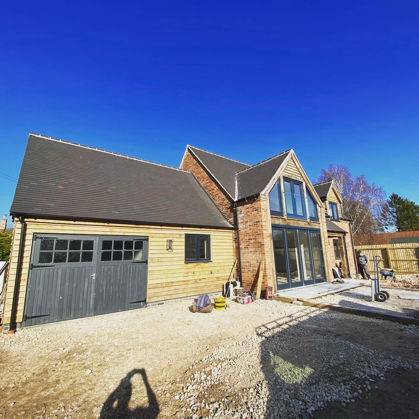 Newly built house with brick and wooden siding, large glass windows, a garage, and a fenced yard under a clear blue sky.