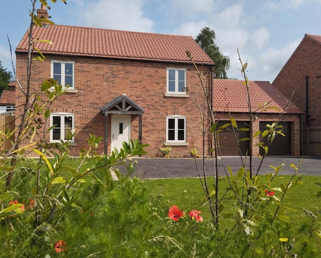 A brick house with a red tiled roof and white-framed windows, surrounded by a green lawn and a flower garden in the front.