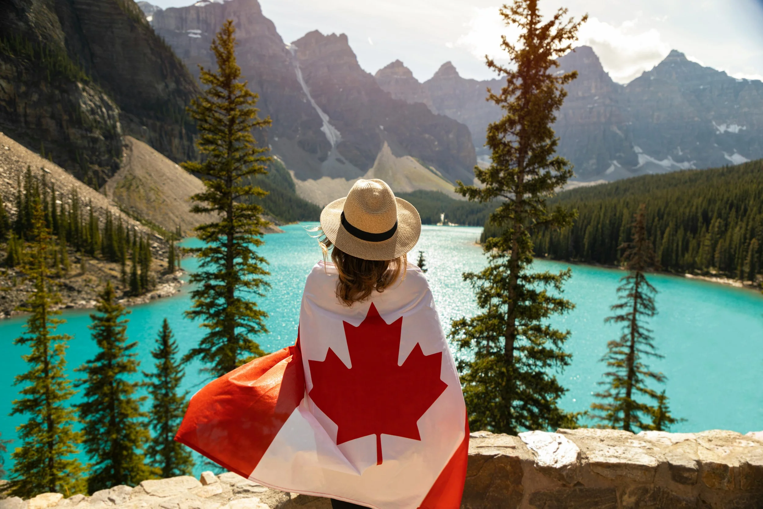 A woman wearing a straw hat and a Canadian flag draped over her shoulders, standing on a stone overlook. She gazes at a turquoise lake surrounded by evergreen trees and rocky mountains under a partly cloudy sky.