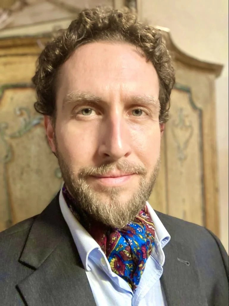 A man with curly hair and a beard smiling, dressed in a suit with a colorful scarf, standing in front of a decorative wooden backdrop.