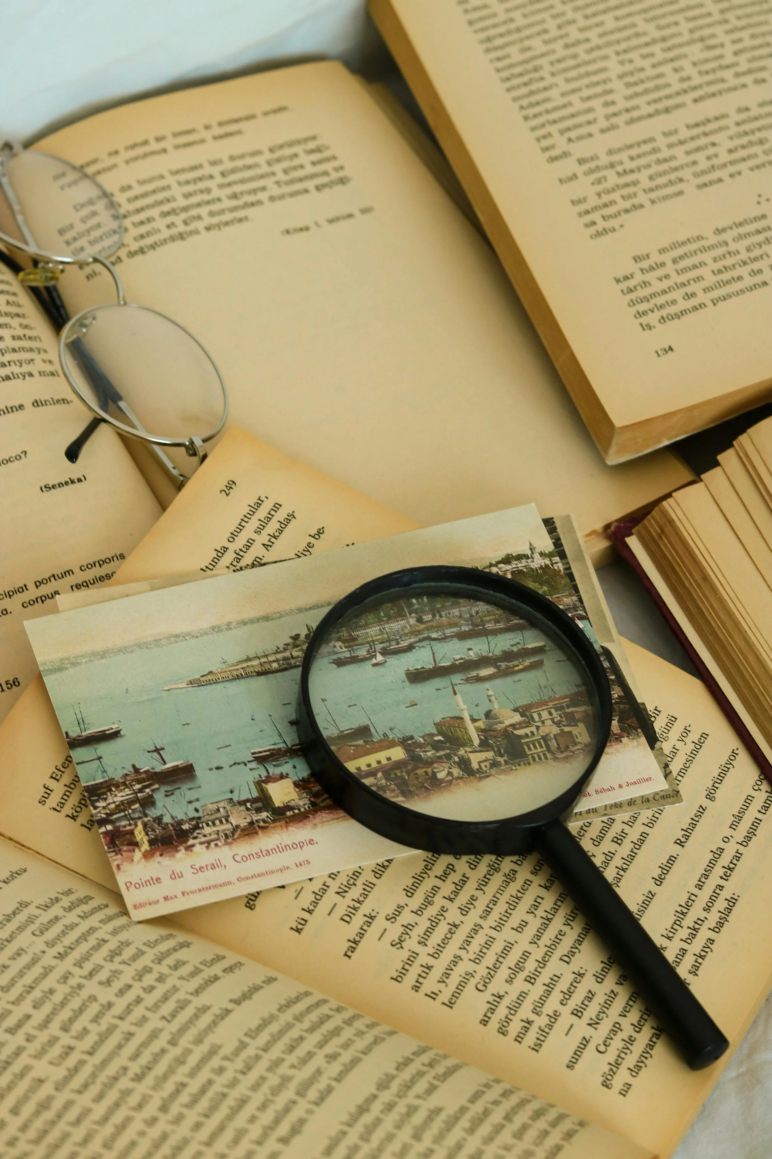 Open books, a pair of glasses, a black magnifying glass, and a postcard of the harbor in Constantinople, Turkey, on a table.