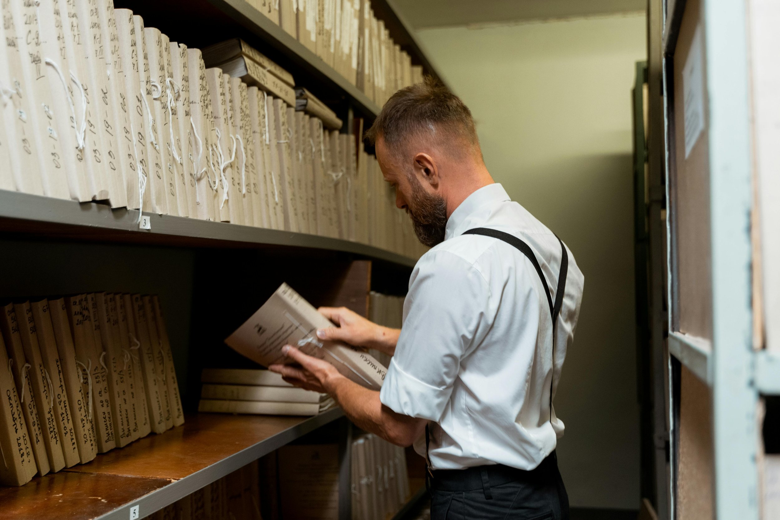 A man with a beard wearing a white shirt and black suspenders looking at a document in an archive or library with shelves of folders and files.