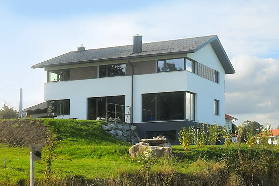 Ein modernes, zweistöckiges Haus auf einer hügeligen Grünfläche mit großen Fenstern und einfachem Design.