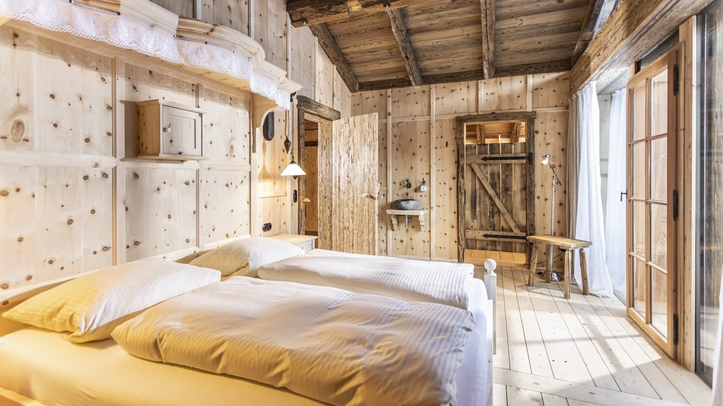 Rustikale Holzzimmer mit Doppelbett, Holzvertäfelung, Holzdecke, Holzfußboden, offener Schrank, Fenster mit weißen Vorhängen, kleines Regal mit Waschbecken, Holzsitzbank, Stehlampe, Tür zu einem Außenbereich.