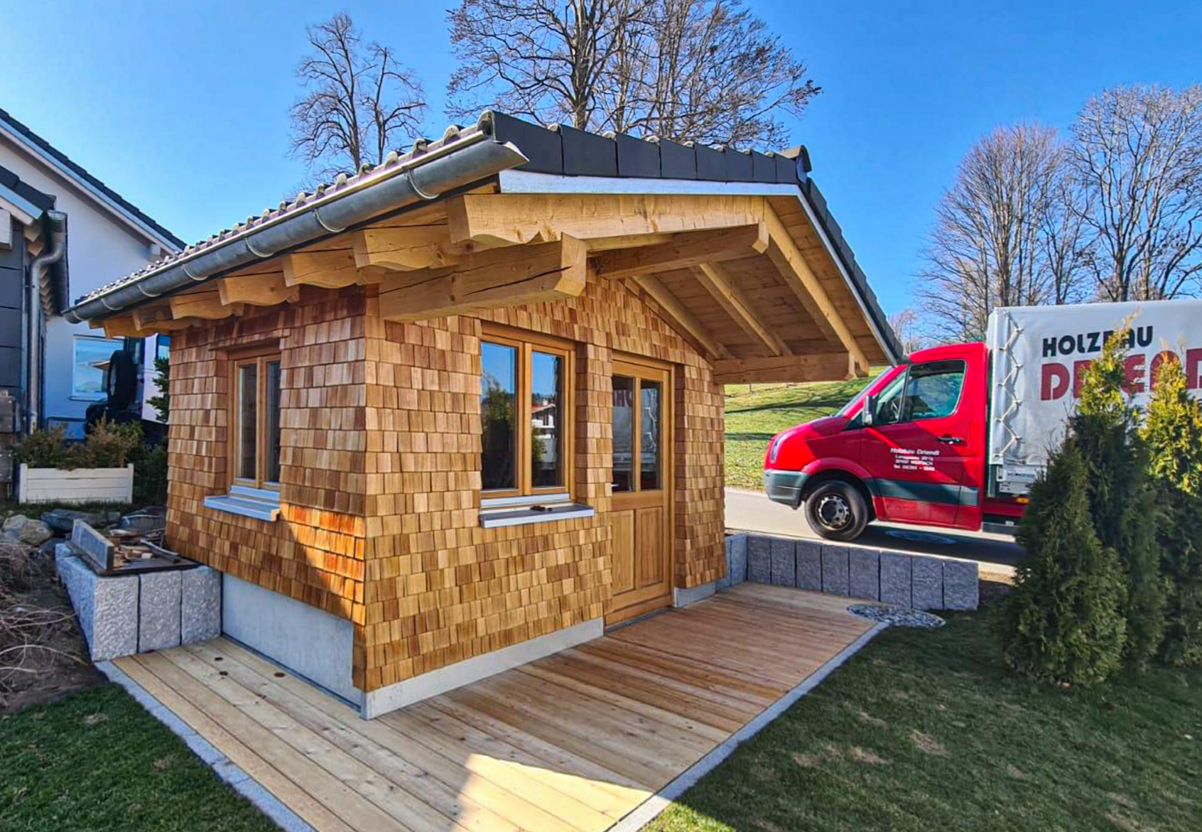 Kleines Holzhaus mit Schindeldach und großen Fenstern auf einem Rasen mit Garten; im Vordergrund eine Holzterrasse; im Hintergrund ein roter Lieferwagen mit Firmenbeschriftung parkend neben dem Haus bei blauem Himmel.