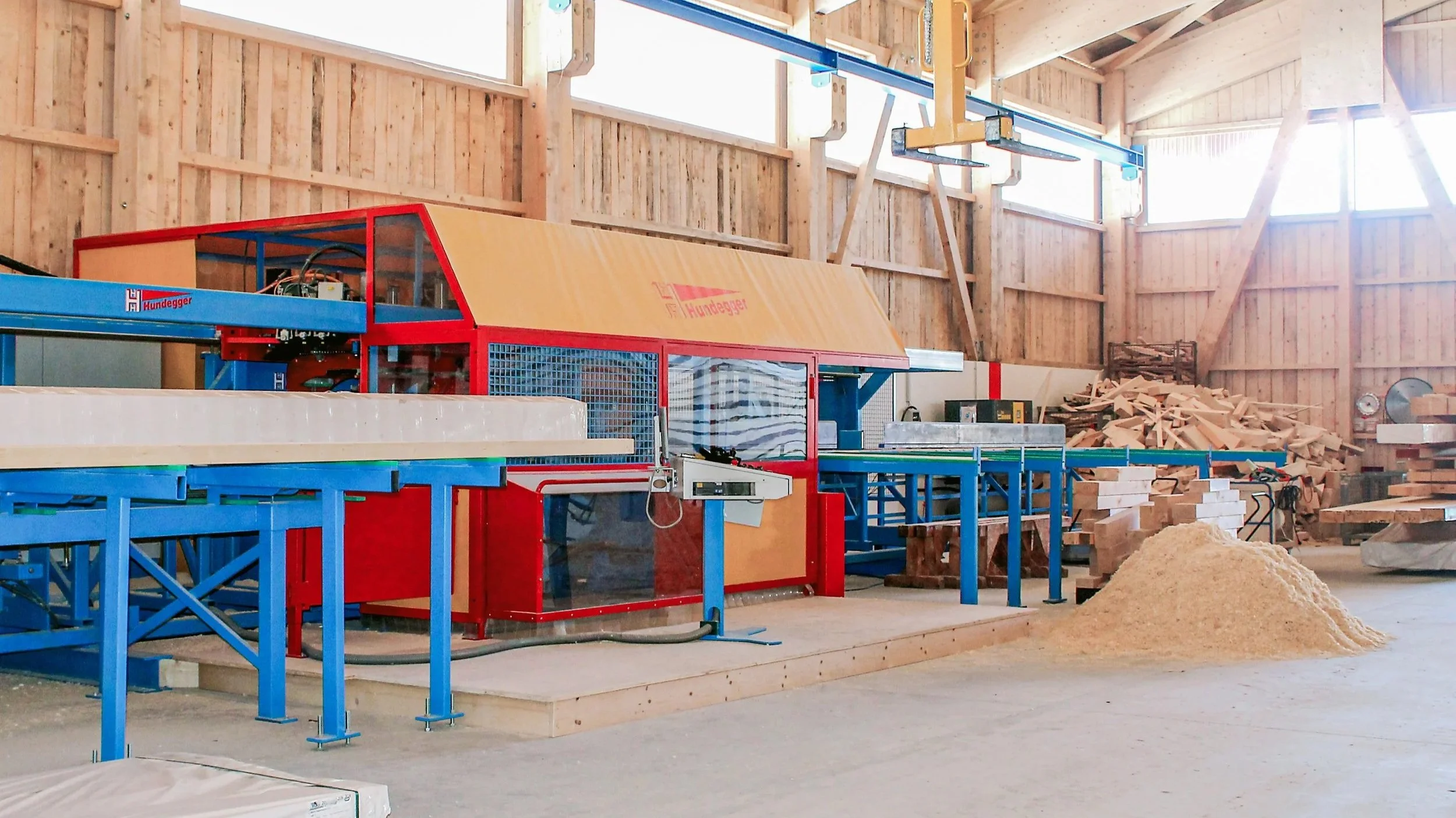 Holzmaschine in einer Werkstatt, umgeben von Holzresten und Holzmaterialien, mit Holzrahmen und großen Fenstern, die Tageslicht hereinlassen.