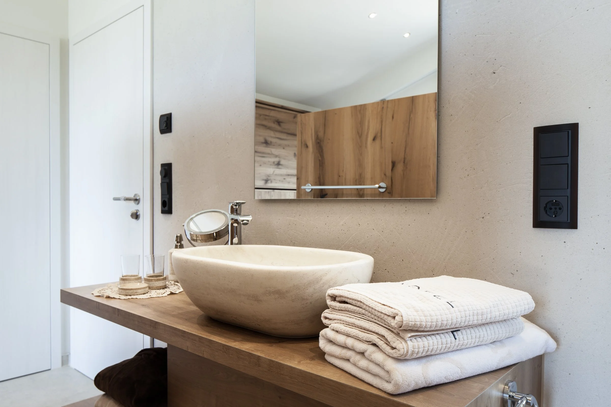 Ein Badezimmer mit einem waschbeckenartigen Waschbecken auf einem Holztisch, darüber ein großer Spiegel, daneben eine weiße Tür, sowie mehrere weiße Handtücher gestapelt.