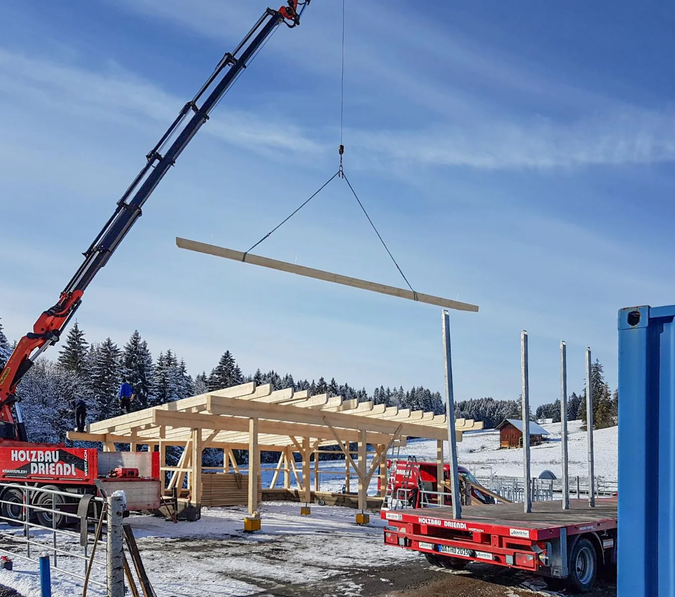 Ein Baukran hebt ein Holzbalken und ist auf einer Baustelle im Schnee. Es wird ein Holzrahmen gebaut, mit Sanddünen, Fichtenbäumen und einem kleinen Haus im Hintergrund.