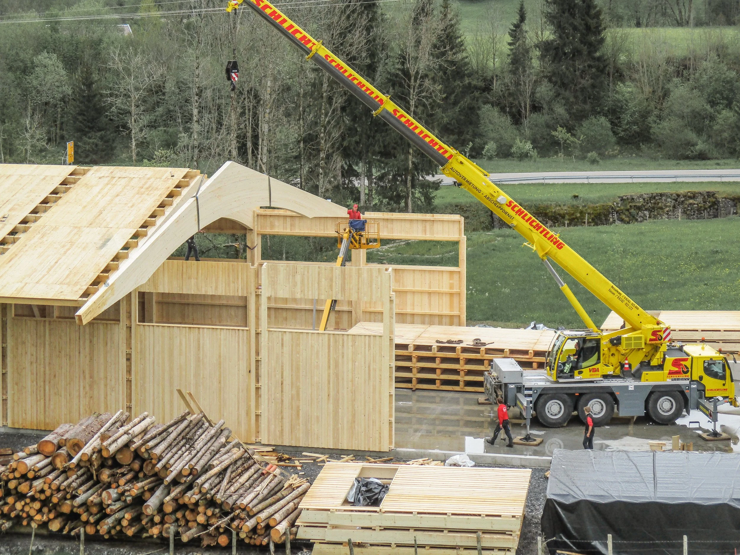 Beim Bau eines Holzhauses wird mit einem Kran Holzstämme gehoben. Bauarbeiter arbeiten an dem Holzrahmen, während viele Baumstämme im Vordergrund lagern.