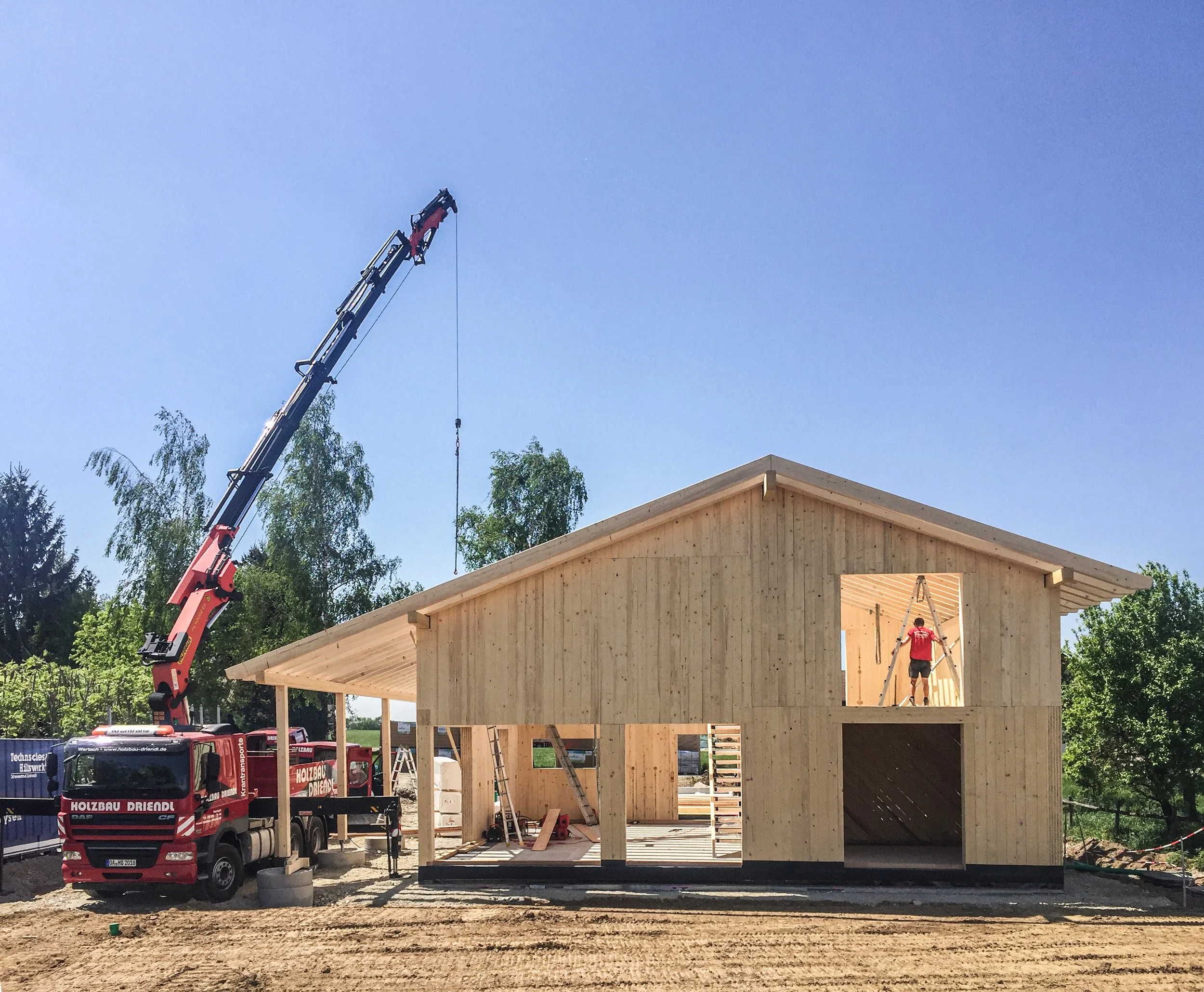 Ein Baukran hebt Baumaterial auf ein Holzhaus im Bau. Ein Arbeiter steht auf einer Leiter im Innenbereich des Hauses.