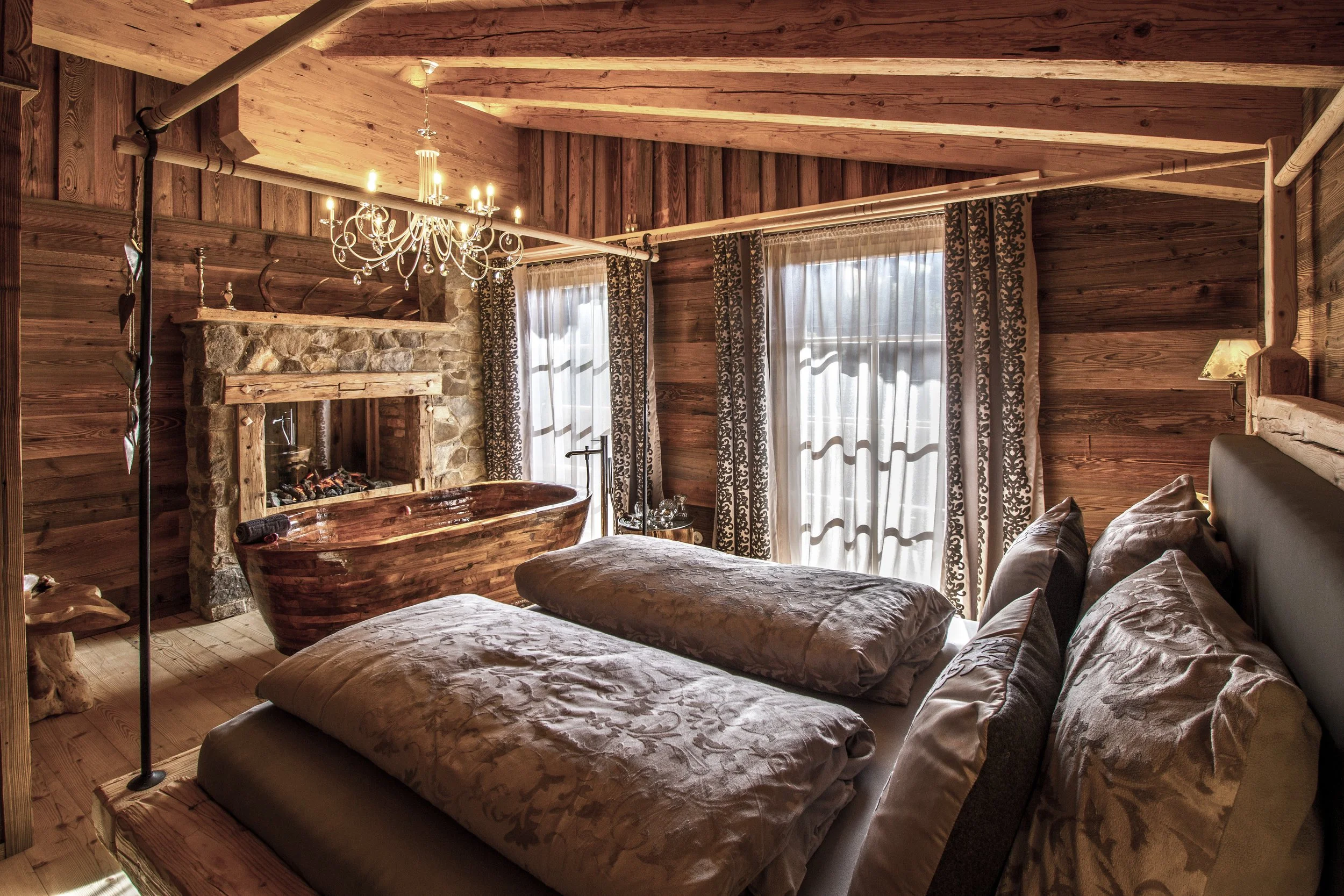 Ein Schlafzimmer mit Holzvertäfelung, einem Kamin, einem alten Badewanne, Bett und Vorhängen vor den Fenstern, dekorativem Kronleuchter an der Decke.