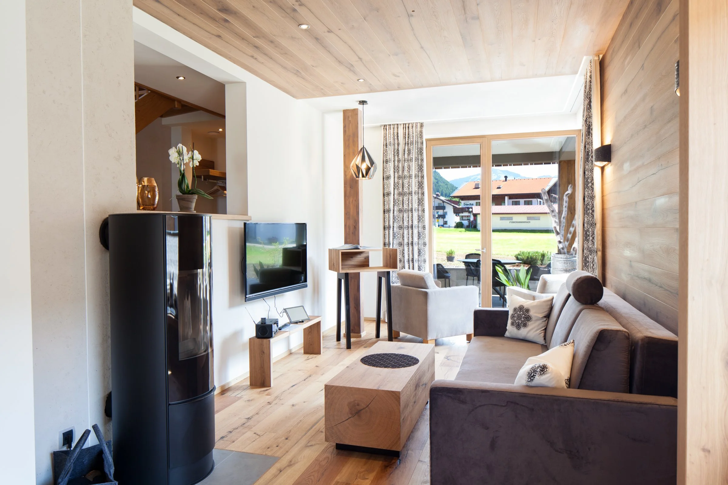 Ein modernes Wohnzimmer mit großen Fenstern, Holzwand und -decken, einem grauen Sofa, Sessel, einem Fernseher und Dekorationsgegenständen. Im Hintergrund sind Berge und Häuser sichtbar.