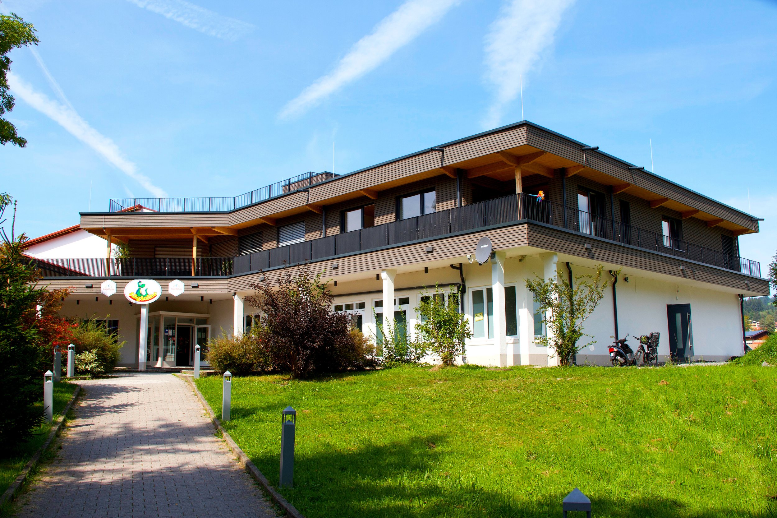 Ein mehrstöckiges Gebäude mit Holzverkleidung und Balkonen, im Vordergrund ein Weg, grüne Rasenflächen, Bäume, Fahrräder und ein blauer Himmel mit Wolken.