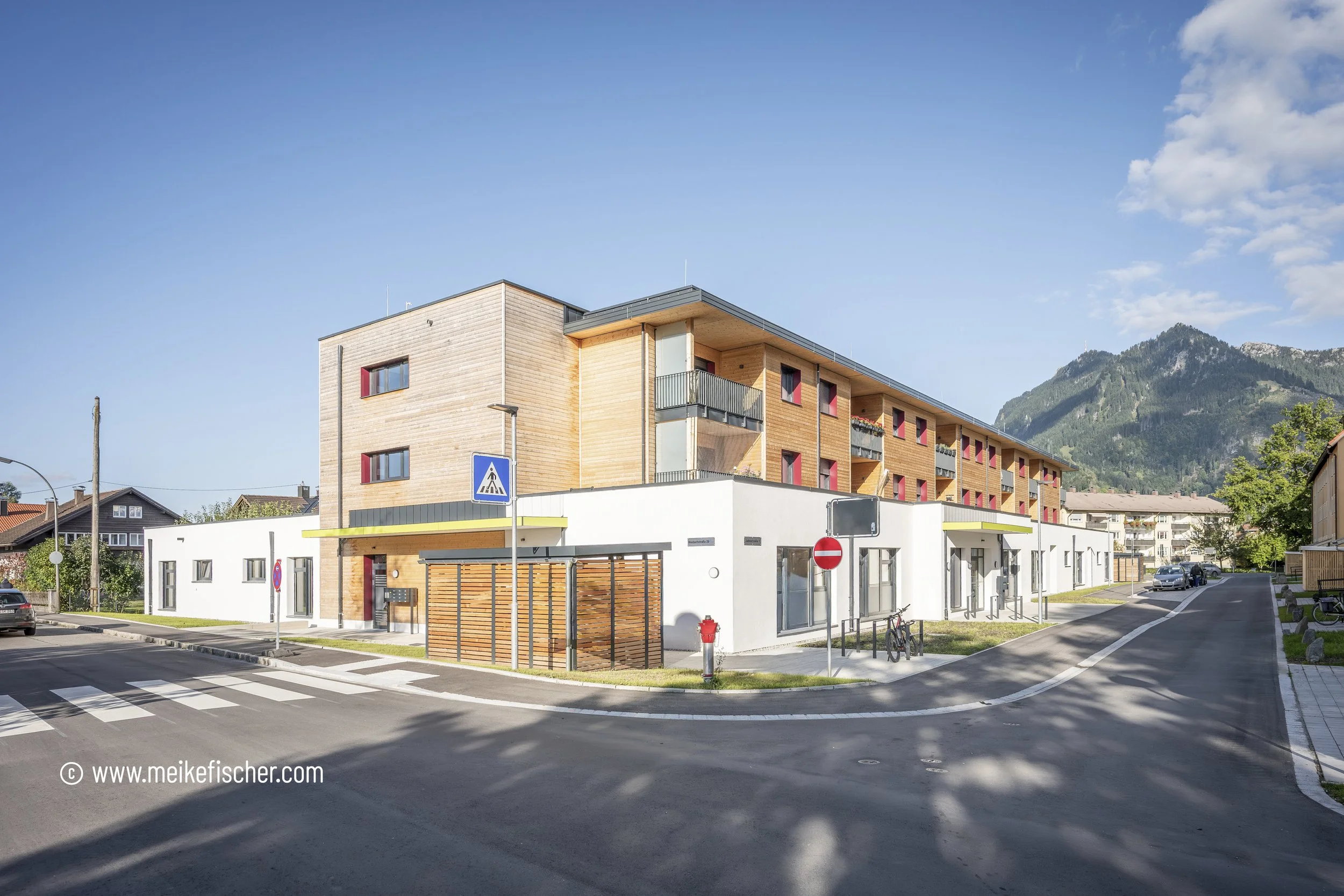 Modernes Mehrfamilienhaus mit Holzelementen im Stadtgebiet, im Hintergrund Berge, blauer Himmel mit wenigen Wolken, Straße mit Fußgängerübergang und parkenden Autos.
