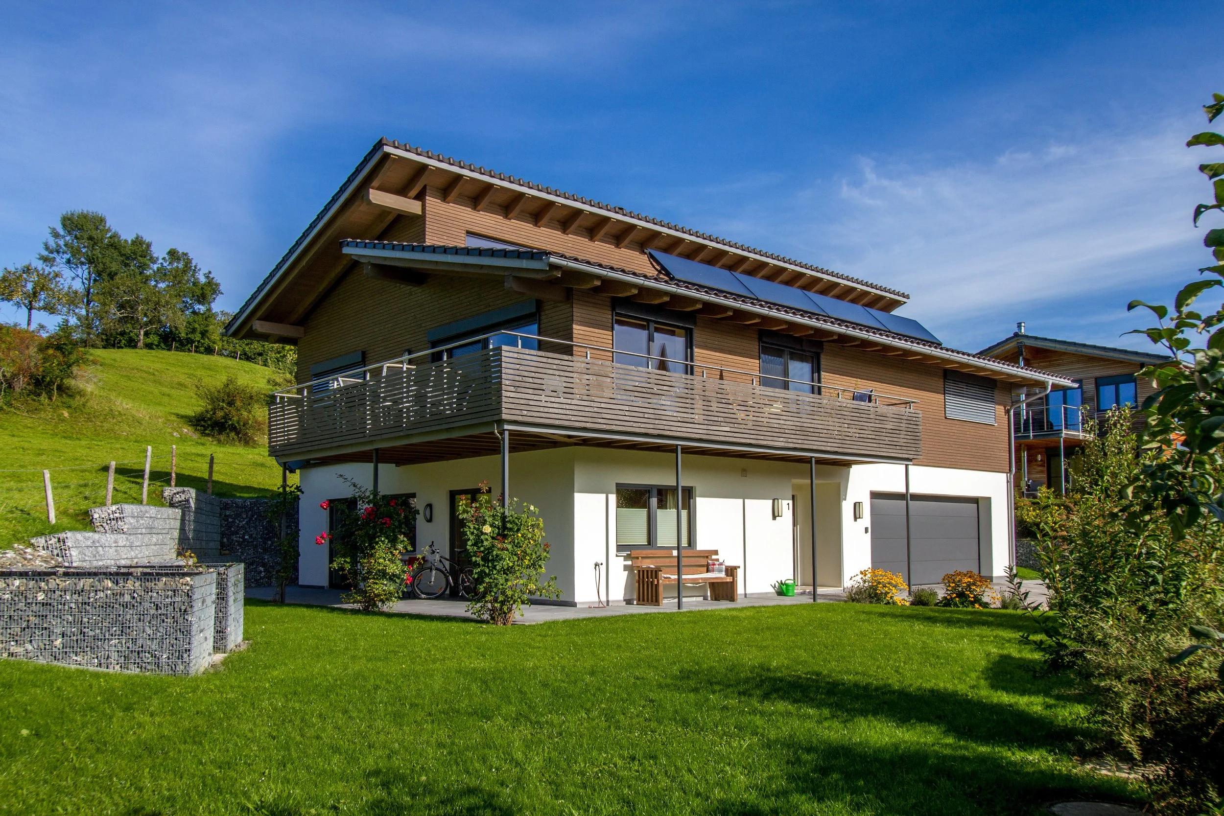 Modernes zweistöckiges Haus mit Balkon, Sonnenkollektoren auf dem Dach, umgeben von grünem Garten und Bäumen bei sonnigem Himmel.