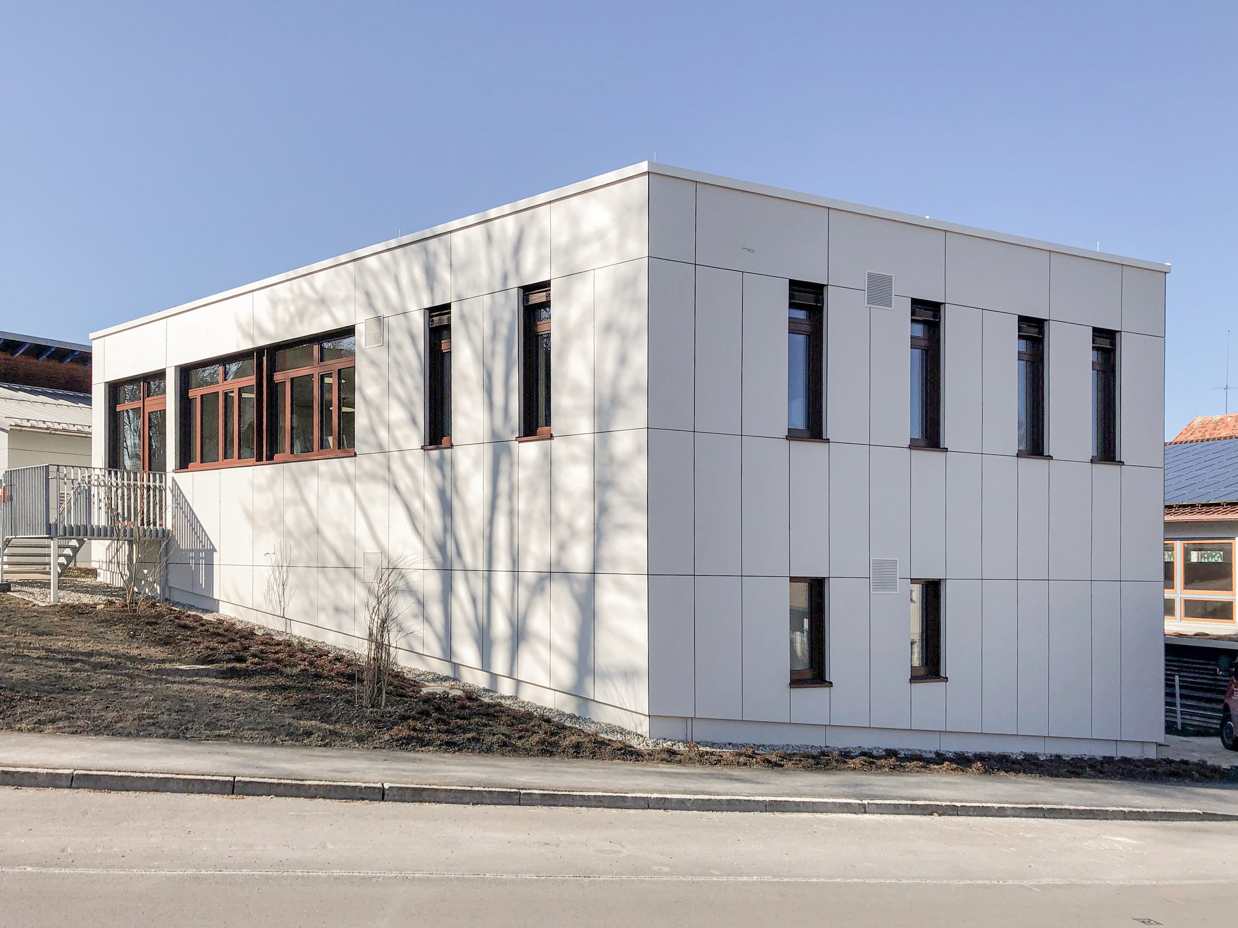 Modernes weißes Gebäude mit großen Fenstern und klaren Linien, im Vordergrund eine Straße und ein Gehweg, einige Bäume ohne Blätter, blauer Himmel.