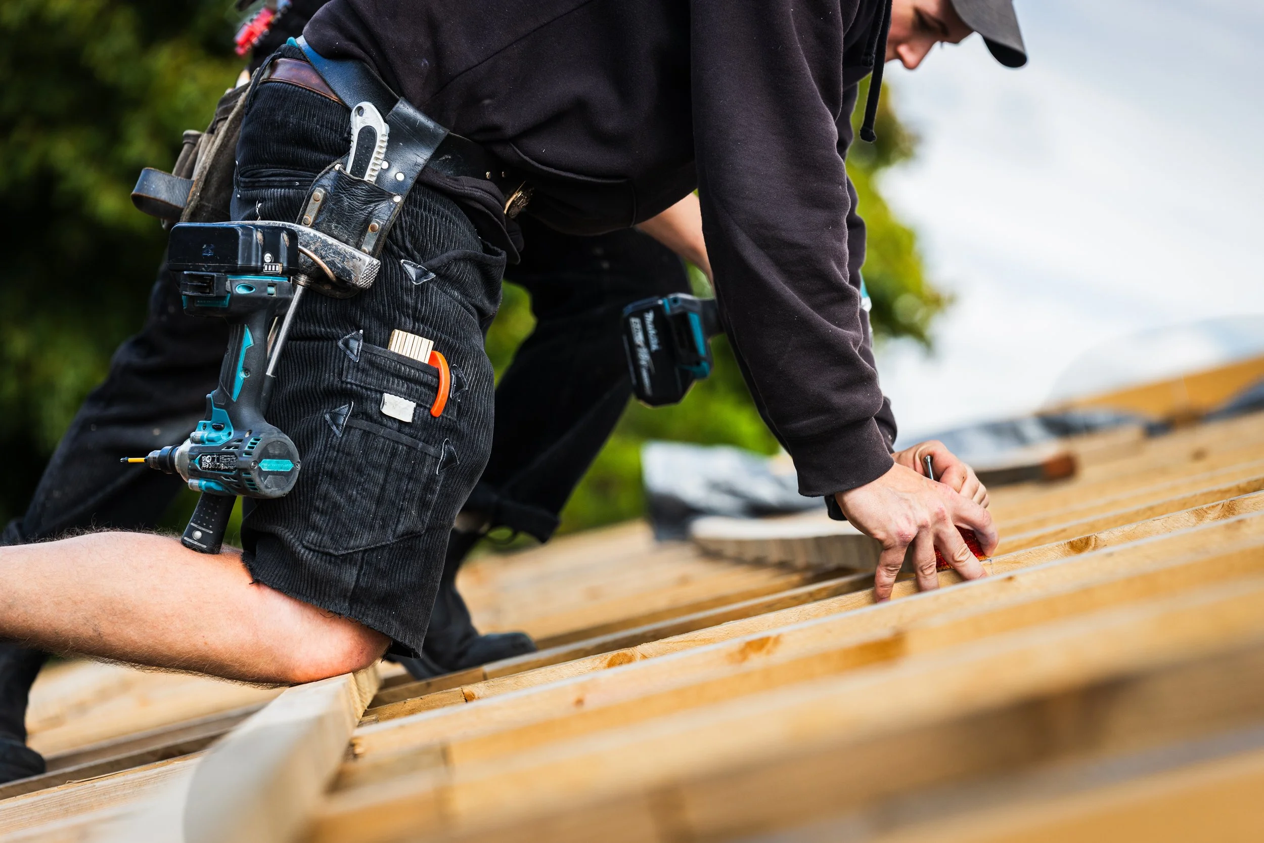 Ein Handwerker arbeitet an einem Holzrahmen im Freien, trägt Werkzeuge in einem Gürtel und verwendet ein Elektrowerkzeug, während er auf dem Boden kniet.