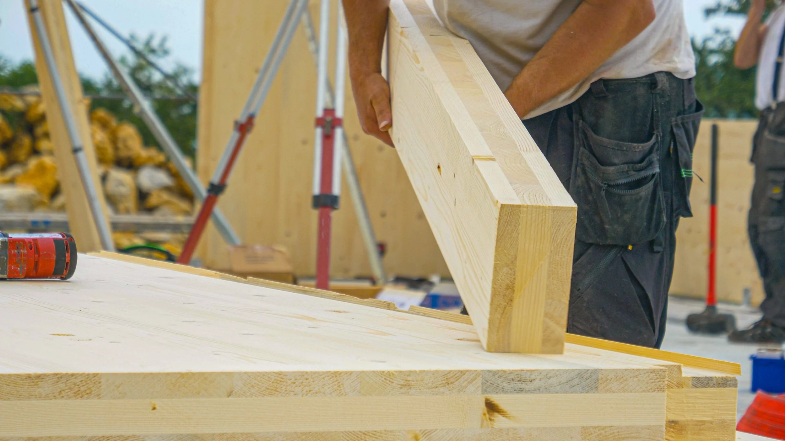 Handwerker arbeitet im Bauwesen, hält ein Stück Holz bei einer Baustelle im Freien, mit Werkzeug und Baumaterial umgeben.