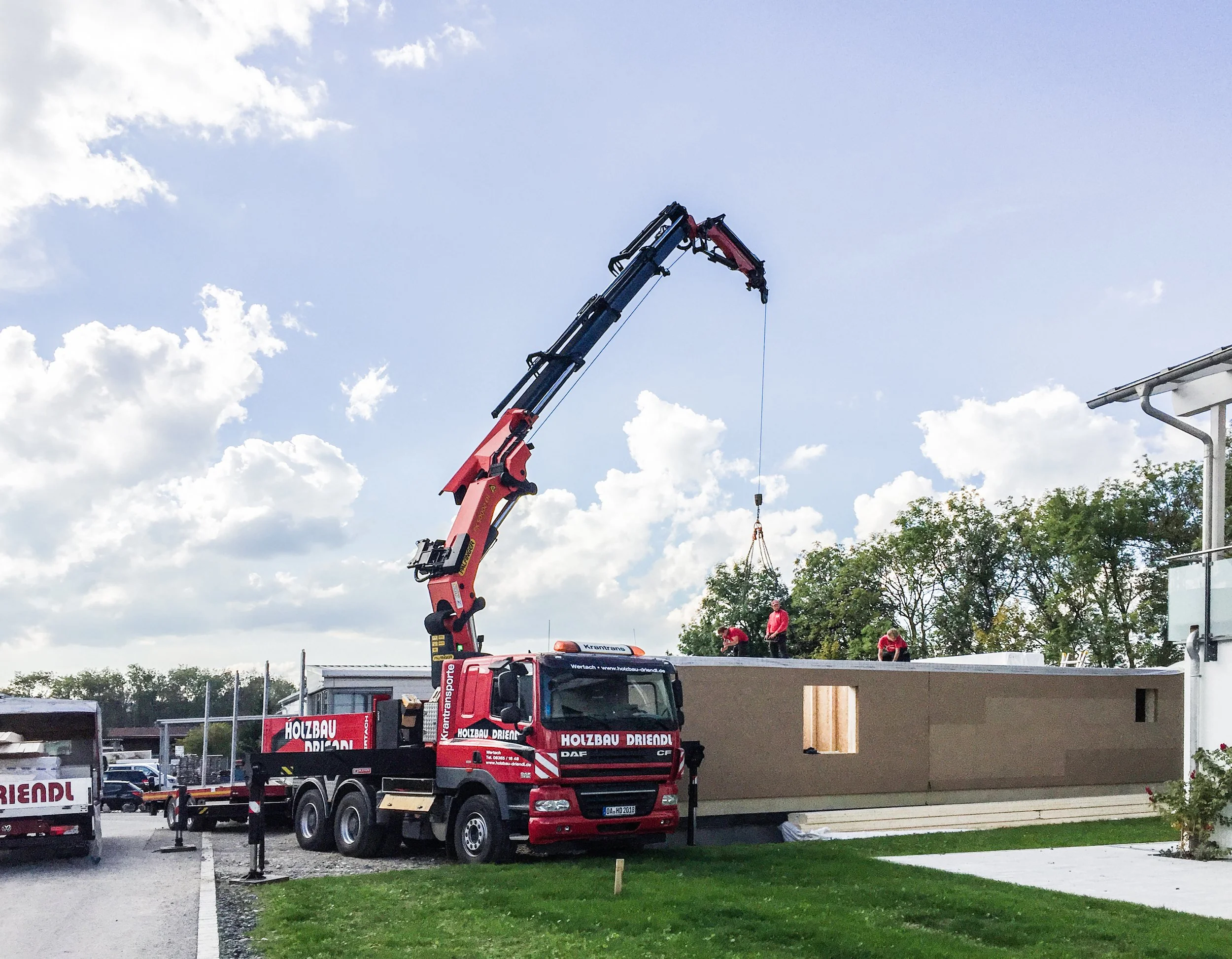 Ein roter Kran hebt Materialien auf ein Gebäude unter Bauarbeiten. Mehrere Arbeiter sind auf dem Bau, einige stehen auf dem Dach. Der Himmel ist teilweise bewölkt.
