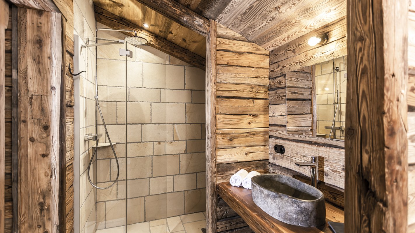 Rustikale Badezimmereinrichtung mit einem offenen Duschbereich auf der linken Seite und einem Waschbecken aus Stein auf der rechten Seite, umgeben von Holzwänden.
