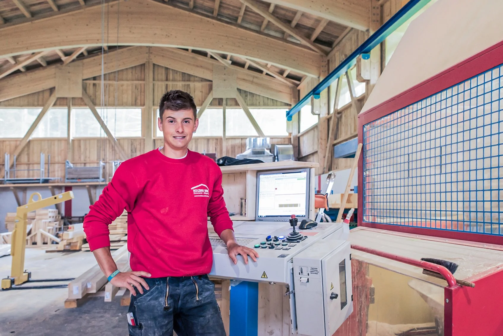 Junger Mann steht in einer Holzwerkstatt neben einer Maschine mit Computersteuerung, im Hintergrund holzverarbeitende Maschinen und Baumaterialien.
