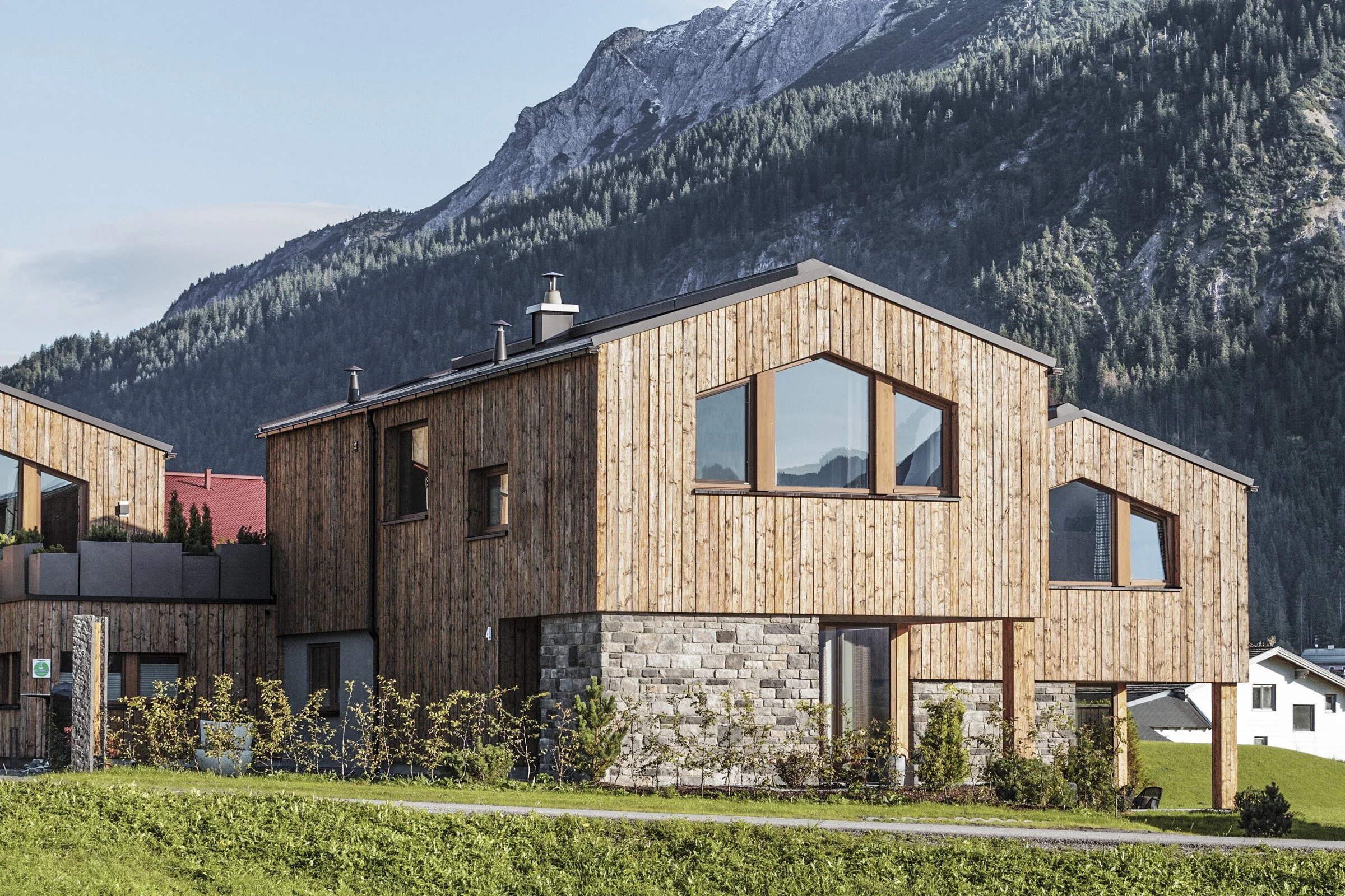 Moderne Holzhäuser vor den Bergen, mit großen Fenstern und Gärten davor.