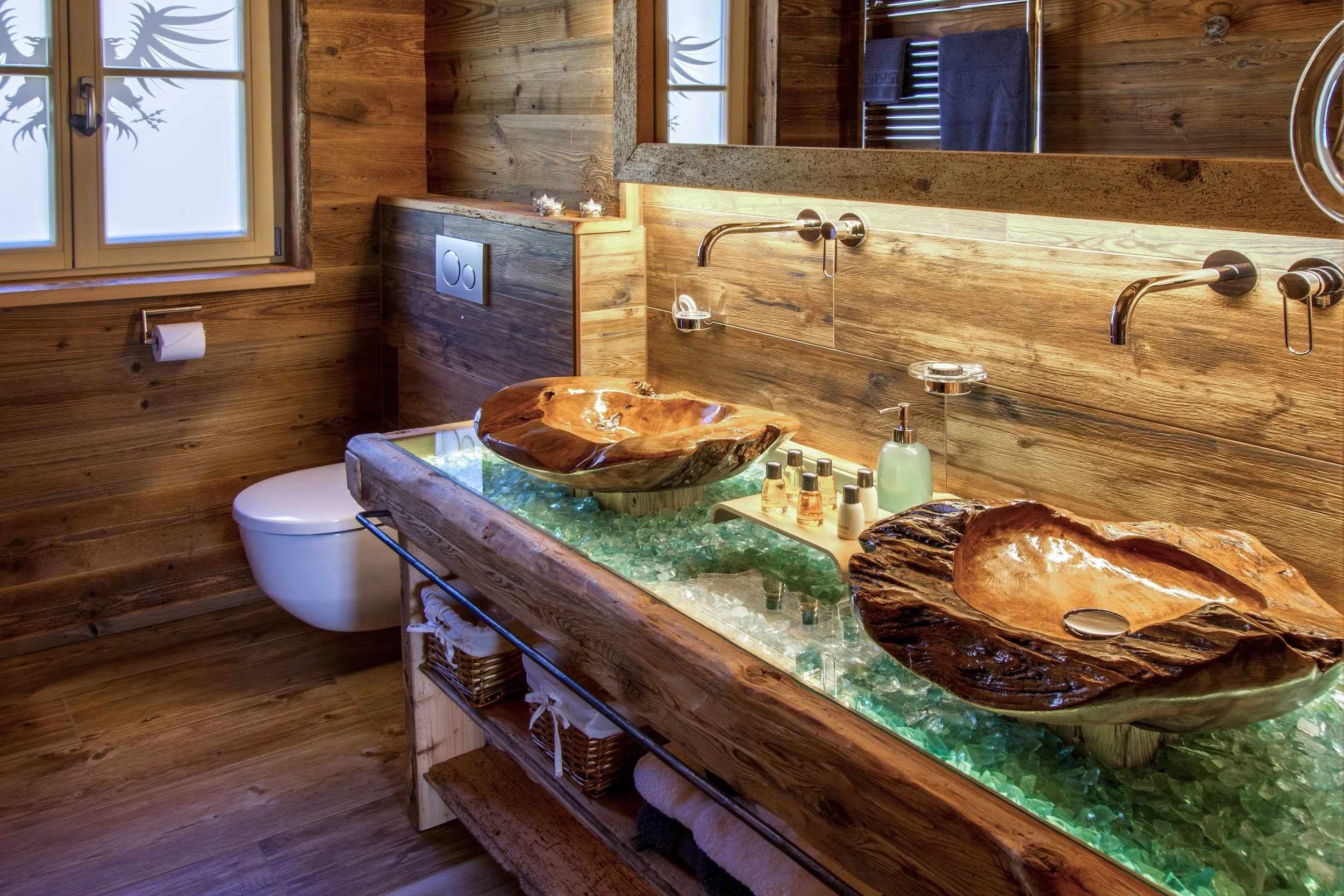 Ein Badezimmer mit Holz verzierten Wänden, einem modernen WC und einem einzigartigen Waschtisch mit einer großen Holzschale als Waschbecken. Das Waschbecken befindet sich auf einem Wasserbecken, das mit Kristallen gefüllt ist, und mehrere kleine Seif