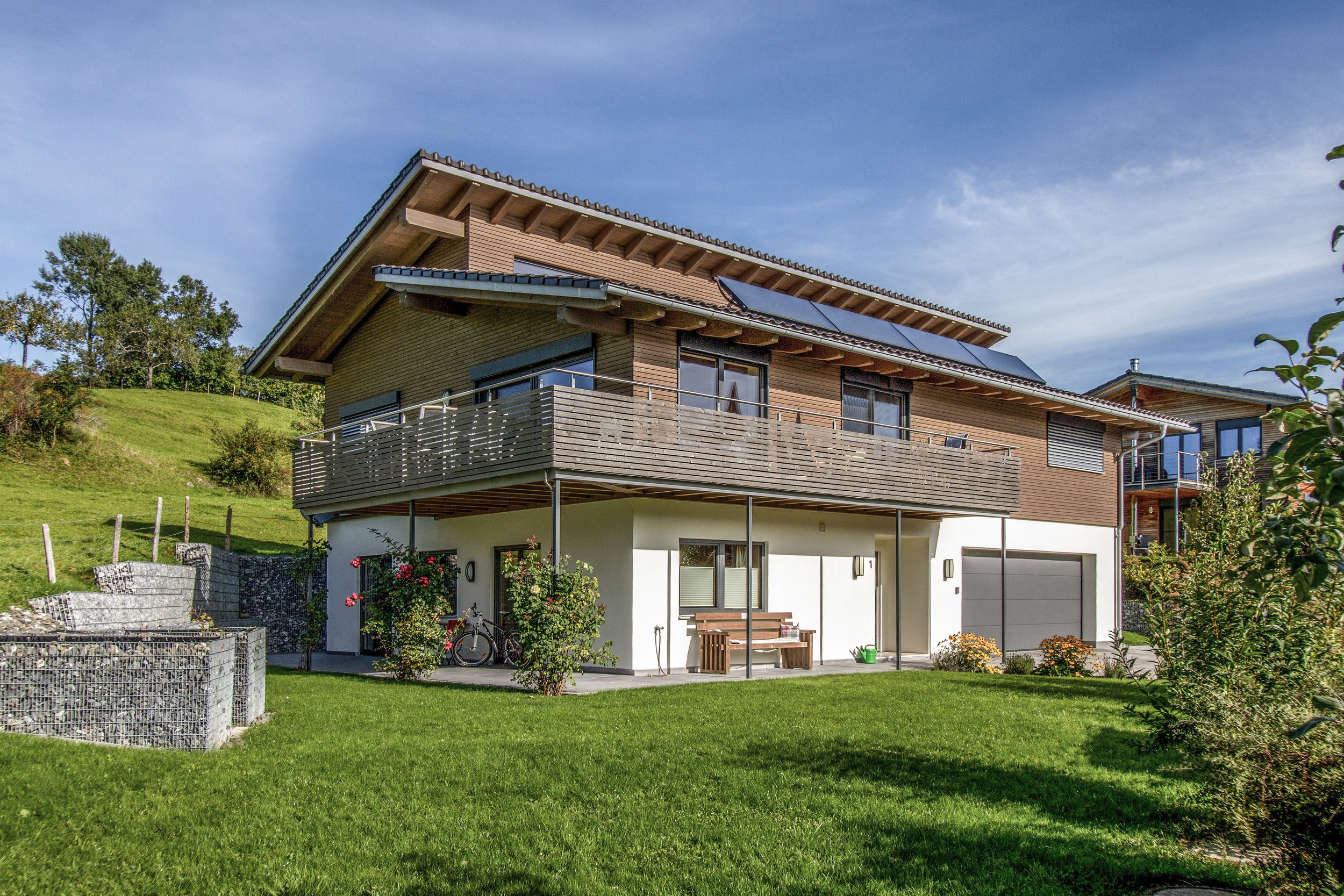 Modern zweistöckiges Haus mit Holzverkleidung, geneigtem Dach mit Solarpanelen, Balkon und Garten mit Blumen und Rasen, umgeben von Bäumen.
